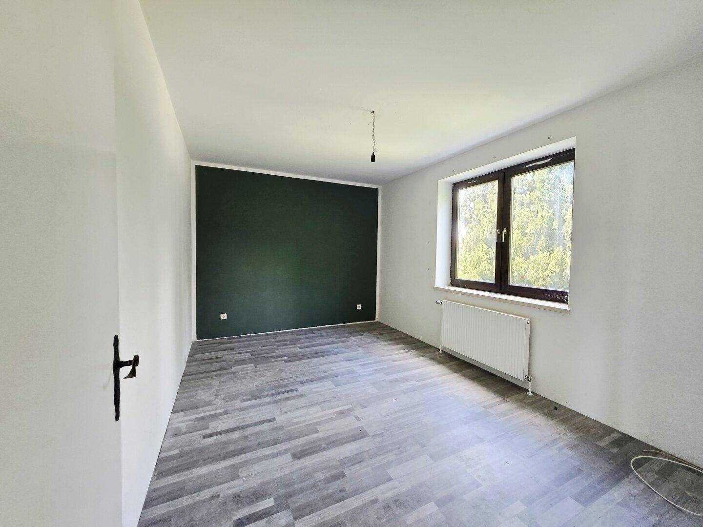 Leeres Schlafzimmer mit grüner Akzentwand, Laminatboden und großem Fenster.