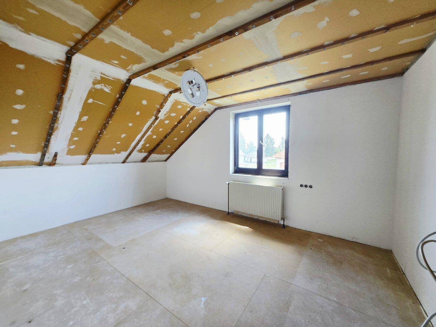 Unfertiger Dachbodenraum mit schrägen Wänden und Fenster, bereit für den Ausbau.