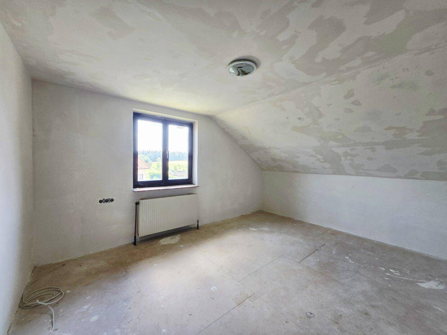 Dachbodenraum im Rohbau mit schrägen Wänden und Fenster, bereit für den Innenausbau.