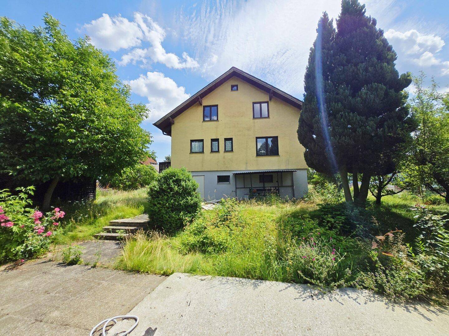 Wohnhausfassade mit Gartenbereich und hohen Bäumen unter einem sonnigen Himmel.