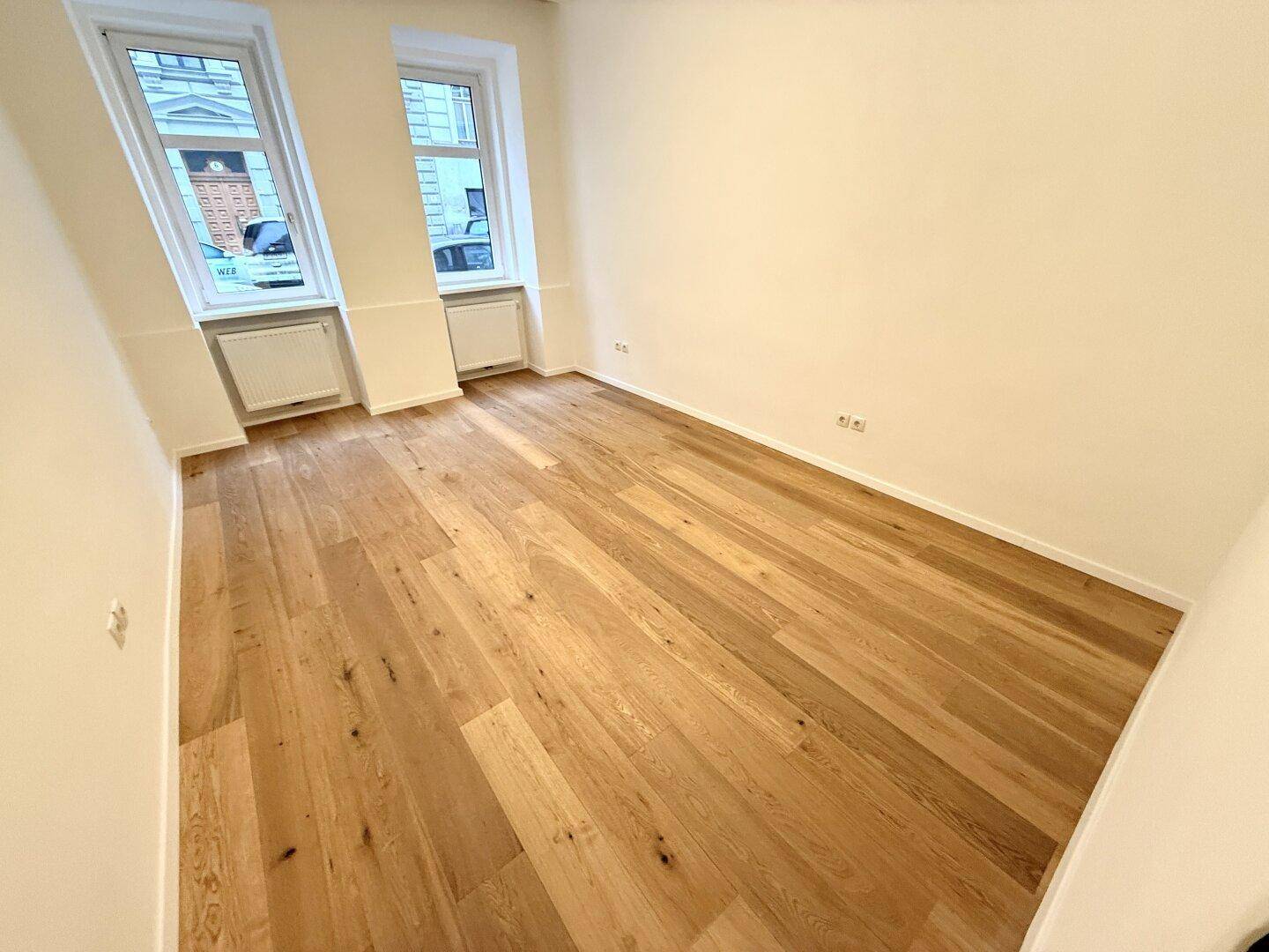 Geräumiges leeres Zimmer mit Holzboden und großen Fenstern, bereit für individuelle Gestaltung und Nutzung.