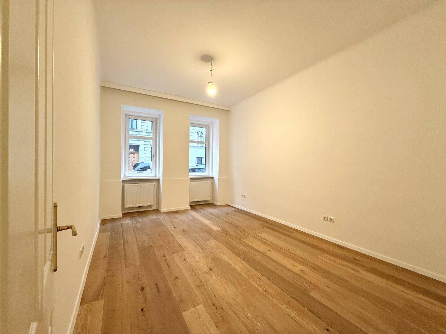 Leeres Zimmer mit hellem Holzboden und zwei Fenstern, bietet viel natürliches Licht und hohe Decken.