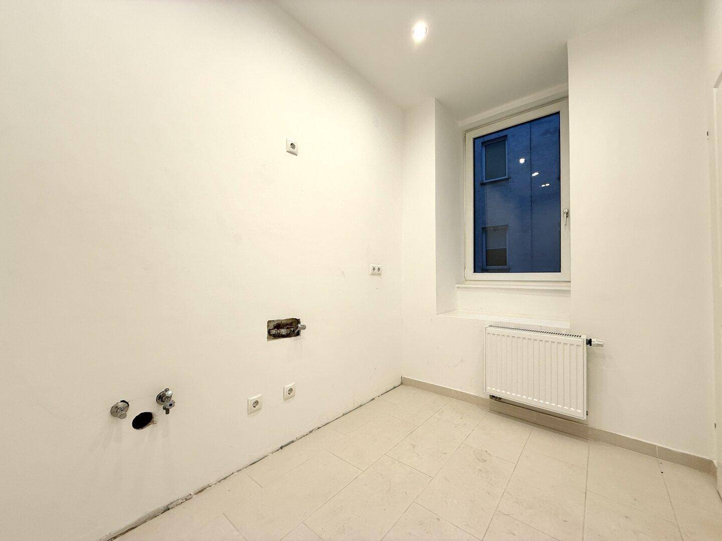 Leerer Raum mit Fliesenboden, Fenster und vorbereiteten Anschlüssen für Sanitärinstallationen.