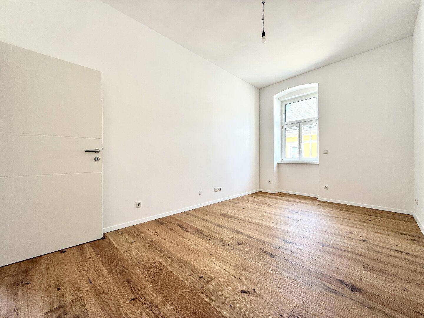 Leeres Zimmer mit Holzboden und einem Fenster, das den Raum mit Tageslicht füllt.