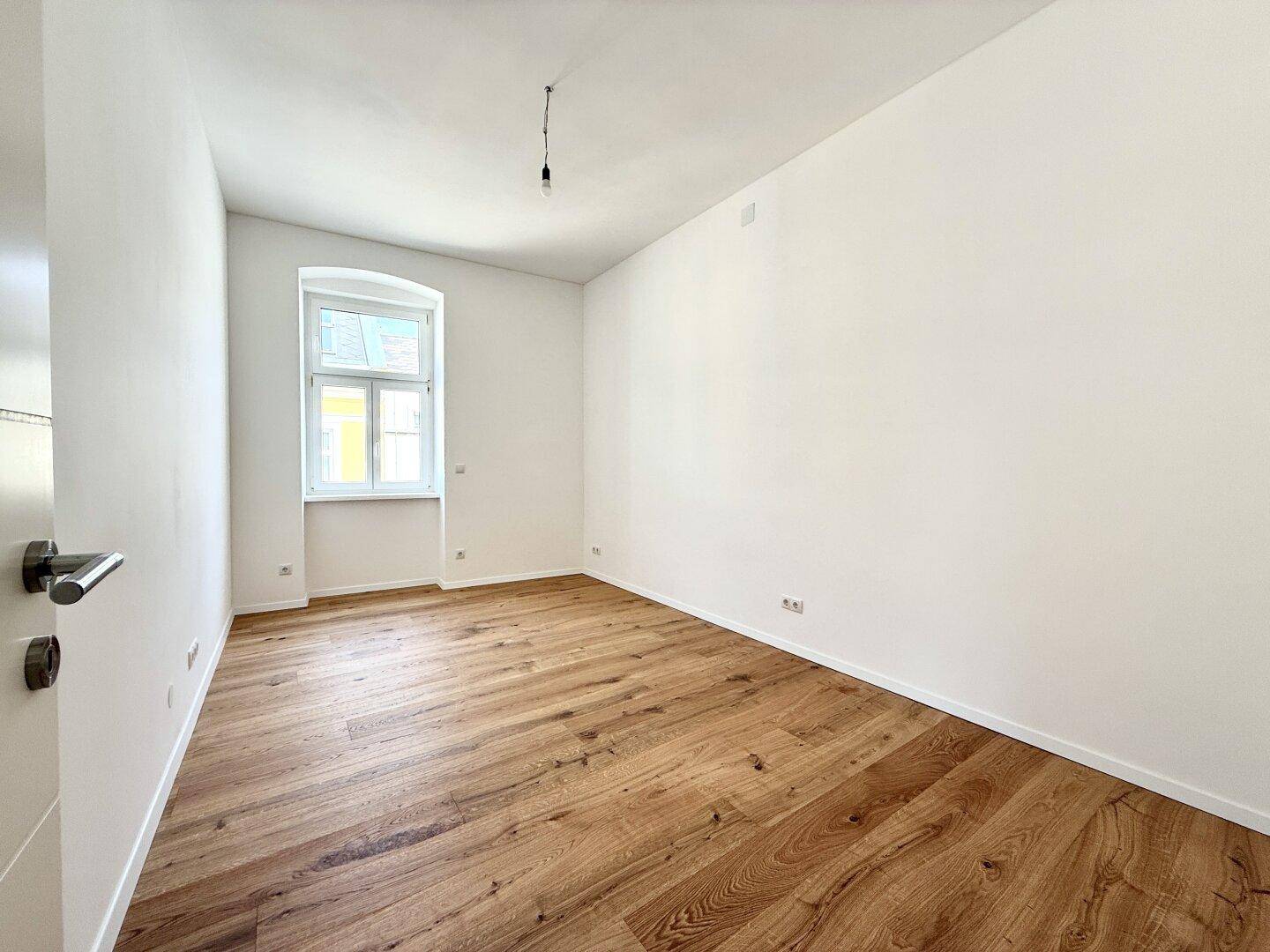 Heller, leerer Raum mit Holzboden und einem Fenster, das viel Licht hereinlässt.