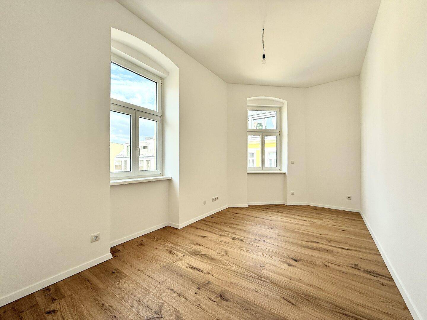 Leeres Zimmer mit Holzboden und zwei Fenstern, die viel natürliches Licht spenden.