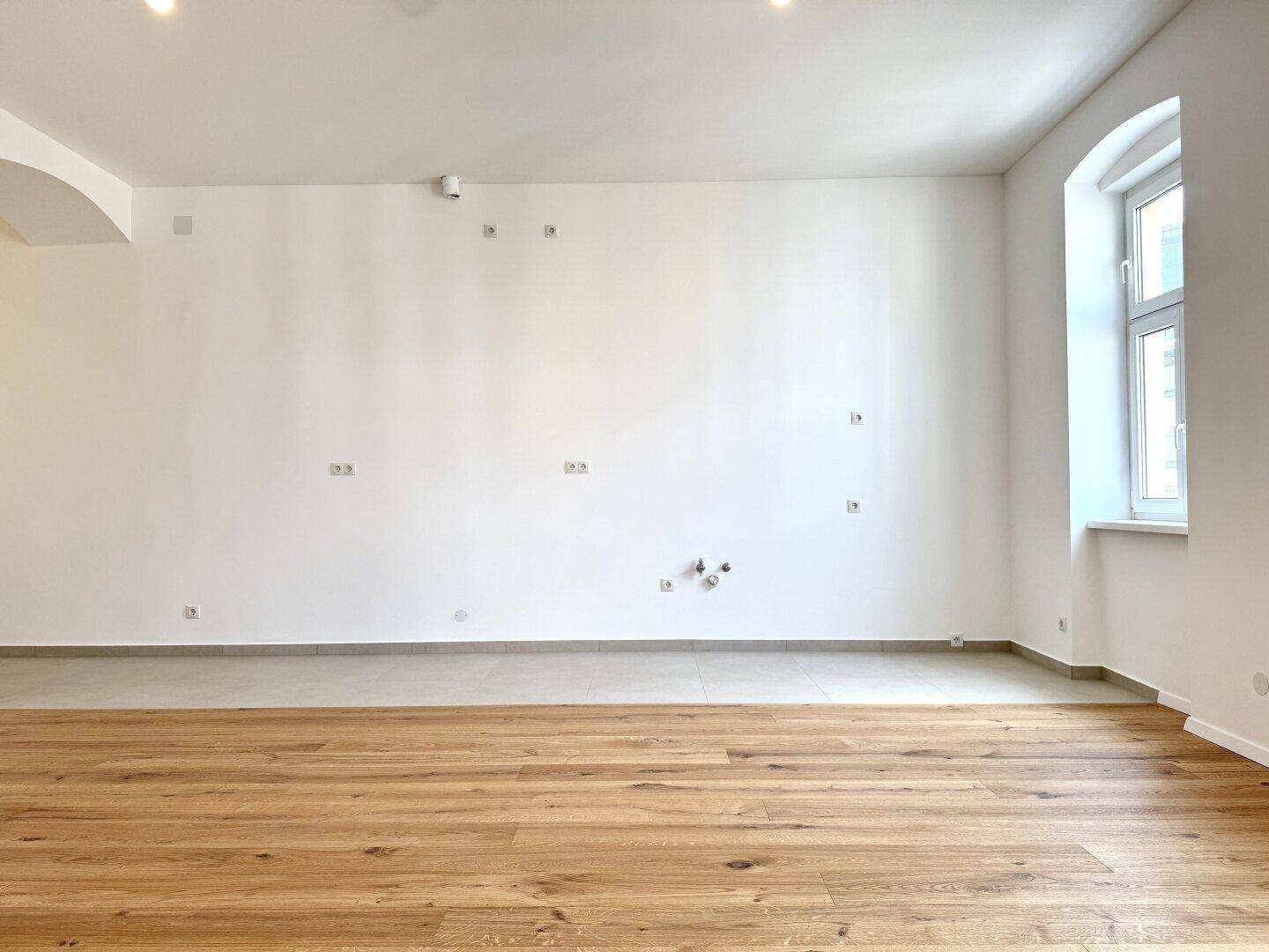 Großer, heller Raum mit Holzboden und einem Fenster, das viel Tageslicht hereinlässt.