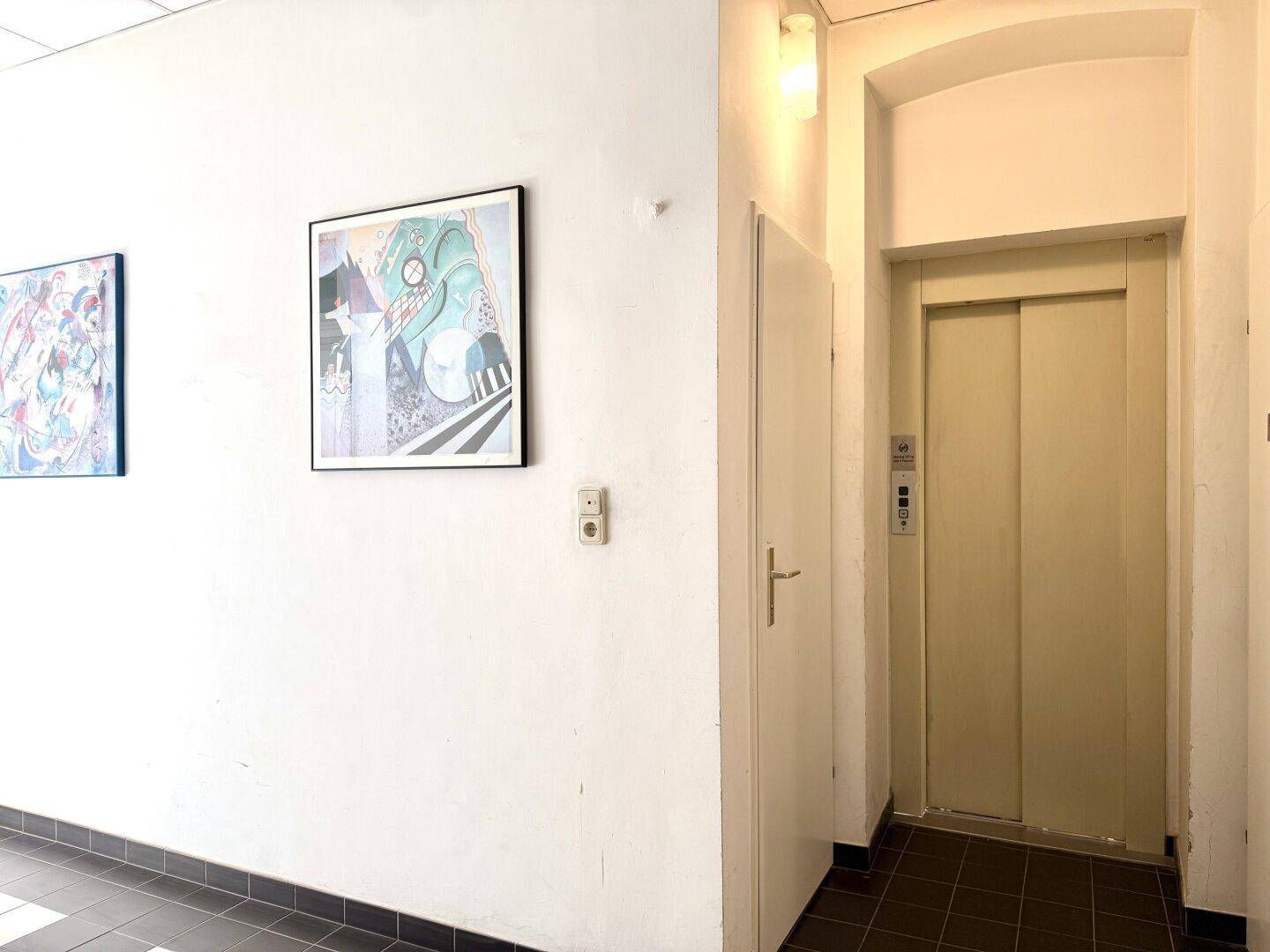 Gemeinschaftsbereich mit Aufzug und Kunstwerken an der Wand, hell und funktional.