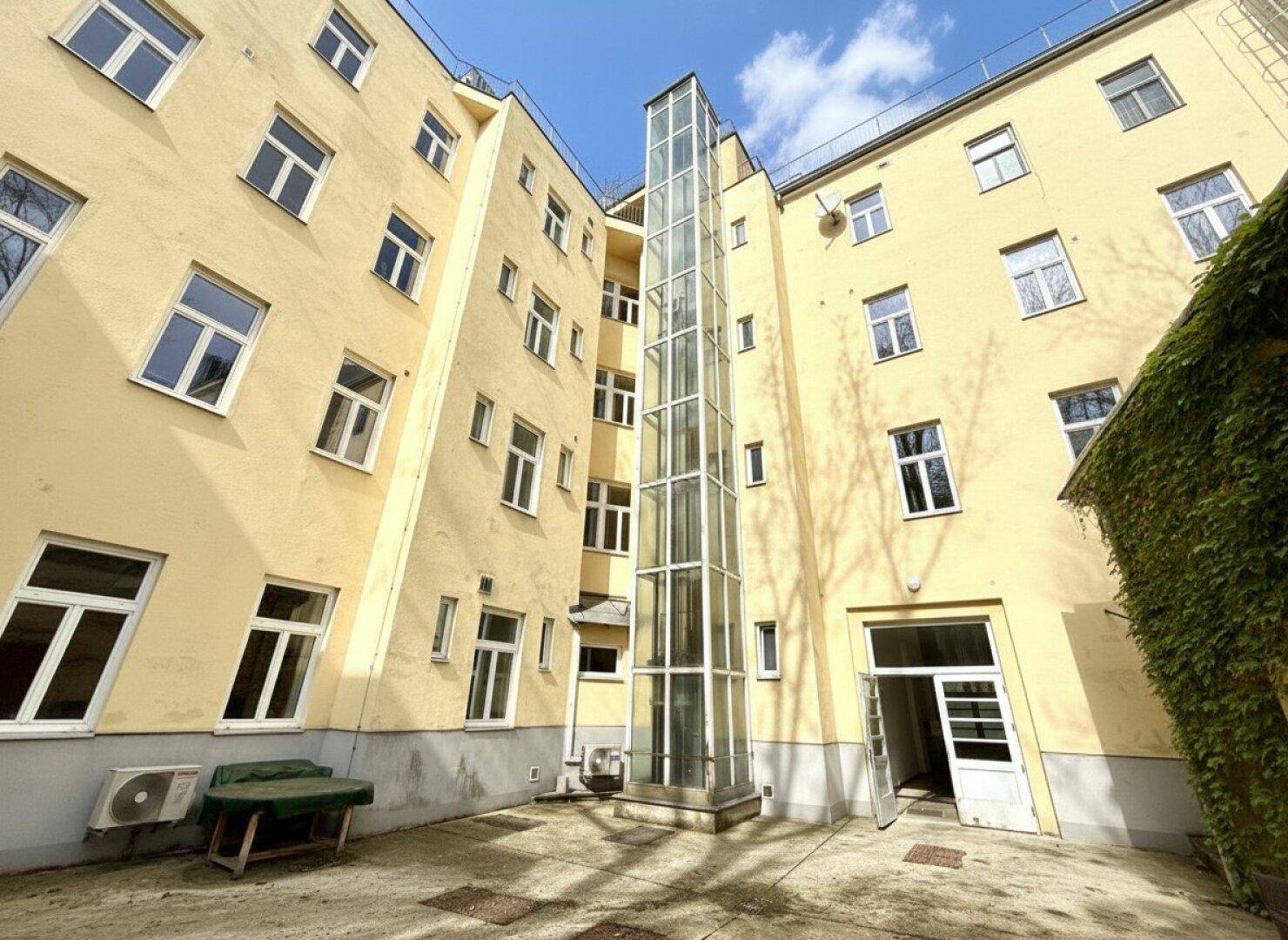 Innenhof mit Glasaufzug und gelber Fassade, umgeben von Wohngebäuden.