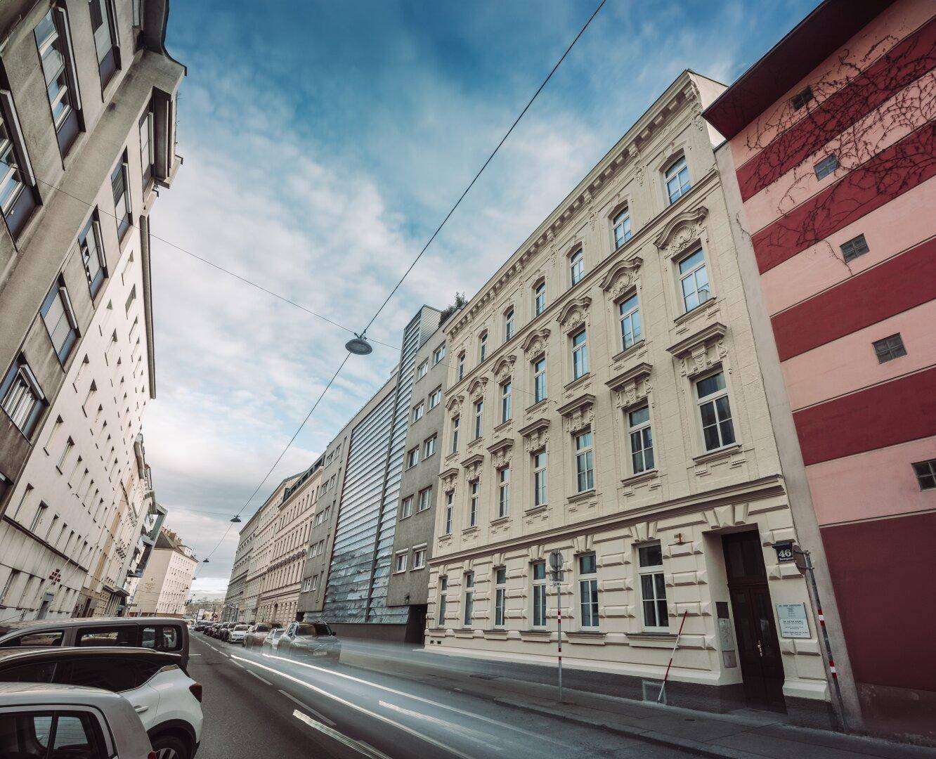 Historisches Gebäude mit klassischer Fassade an einer belebten Stadtstraße unter bewölktem Himmel.