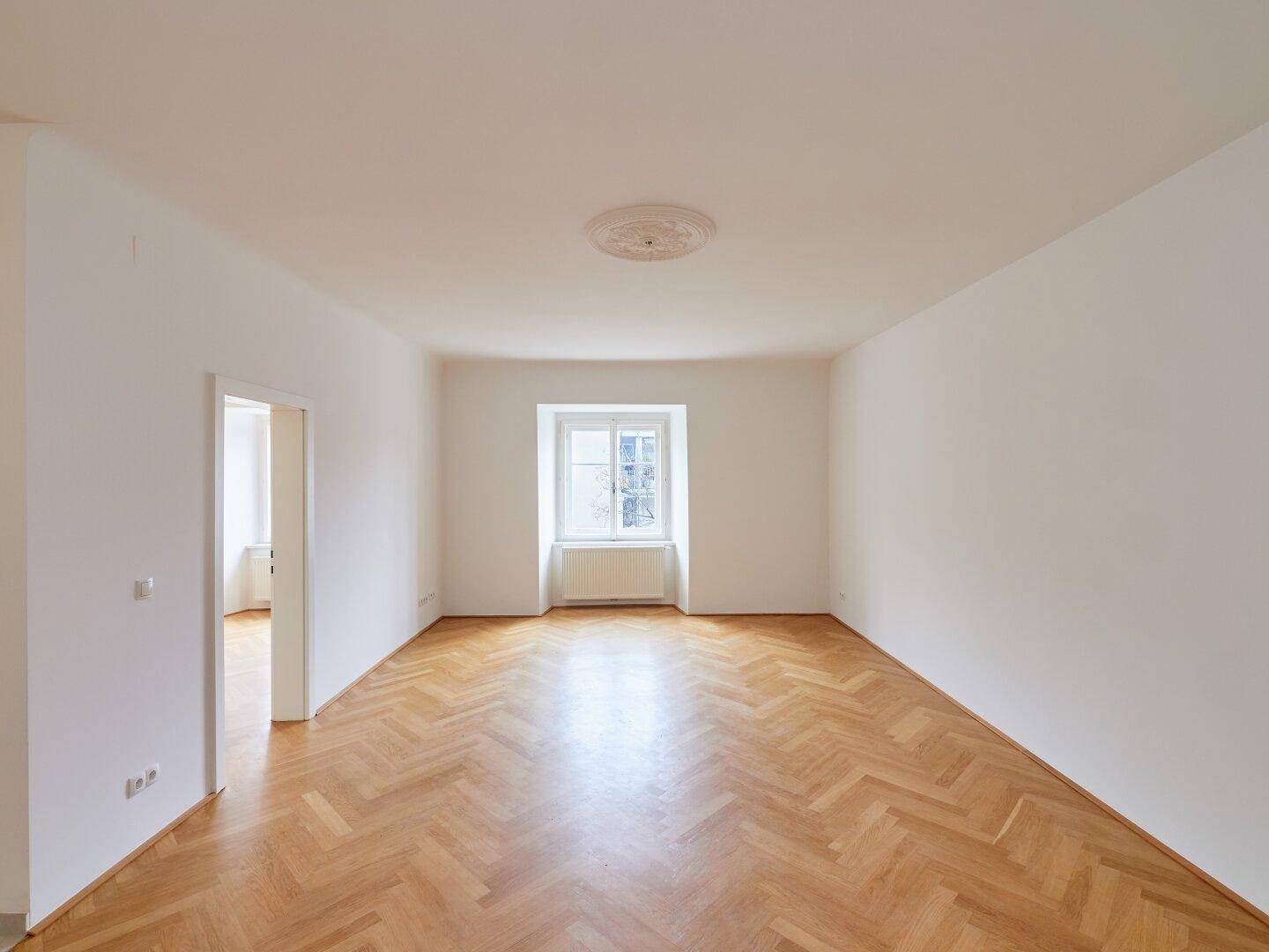 Geräumiges, helles Zimmer mit klassischem Fischgrätparkett und einem Fenster mit Blick ins Grüne.