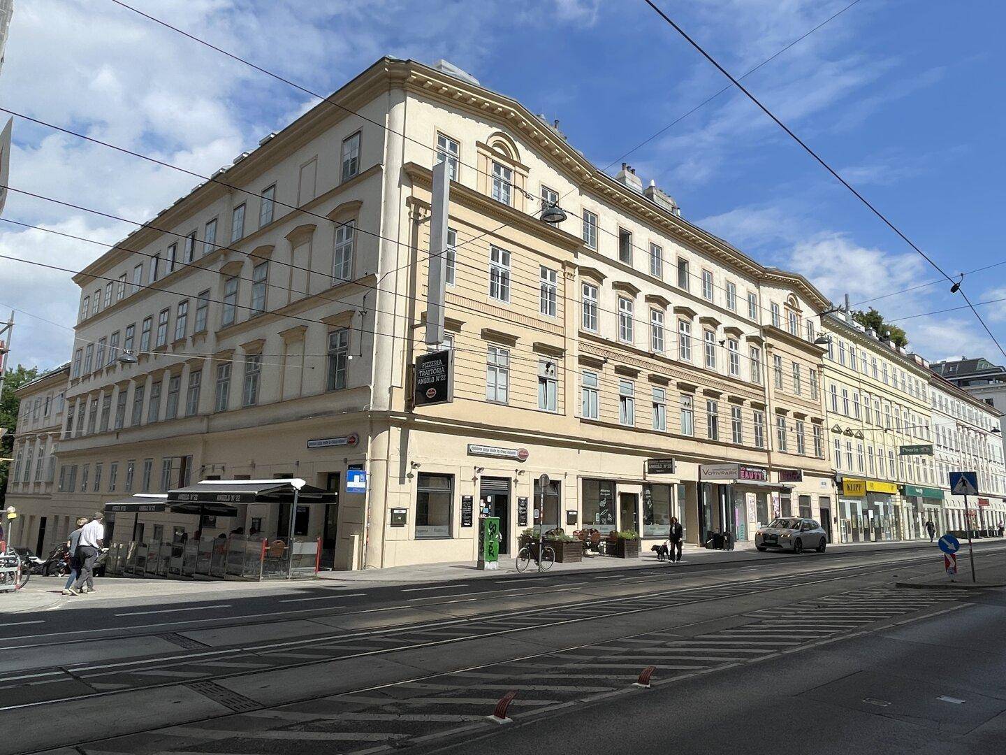 Historisches Eckgebäude mit vielen Fenstern und Geschäften im Erdgeschoss an einer belebten Straße.