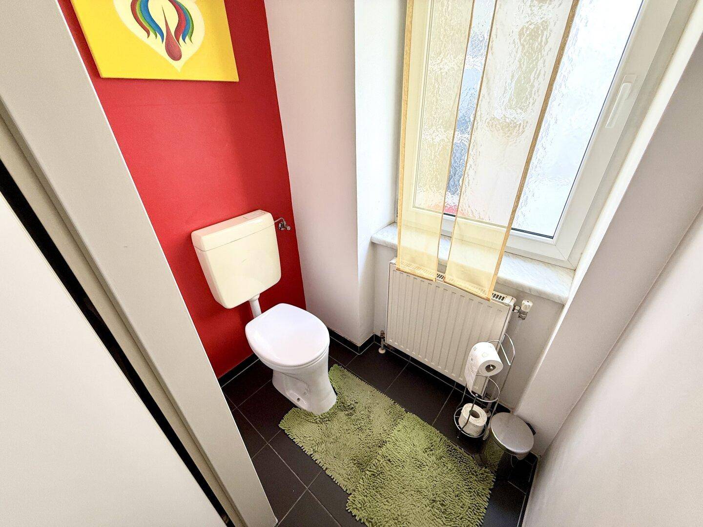 Separate Toilette mit Fenster, dunklen Fliesen und roter Akzentwand, funktional und hell.