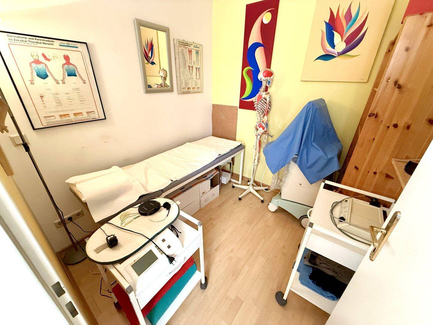 Behandlungsraum mit Liege, medizinischen Geräten und anatomischen Postern, funktional eingerichtet.
