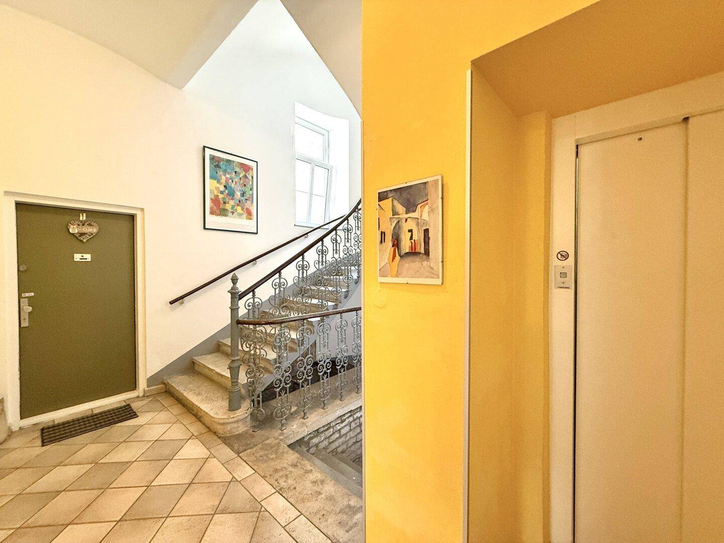 Eingangsbereich mit gefliestem Boden, einer geschwungenen Treppe und einem Aufzug im Hintergrund.