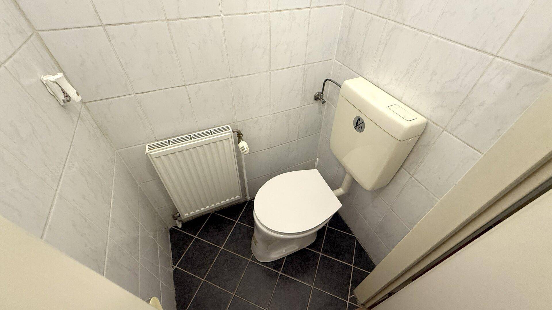 Kleines WC mit weißen Wandfliesen, dunklen Bodenfliesen und einem Heizkörper.