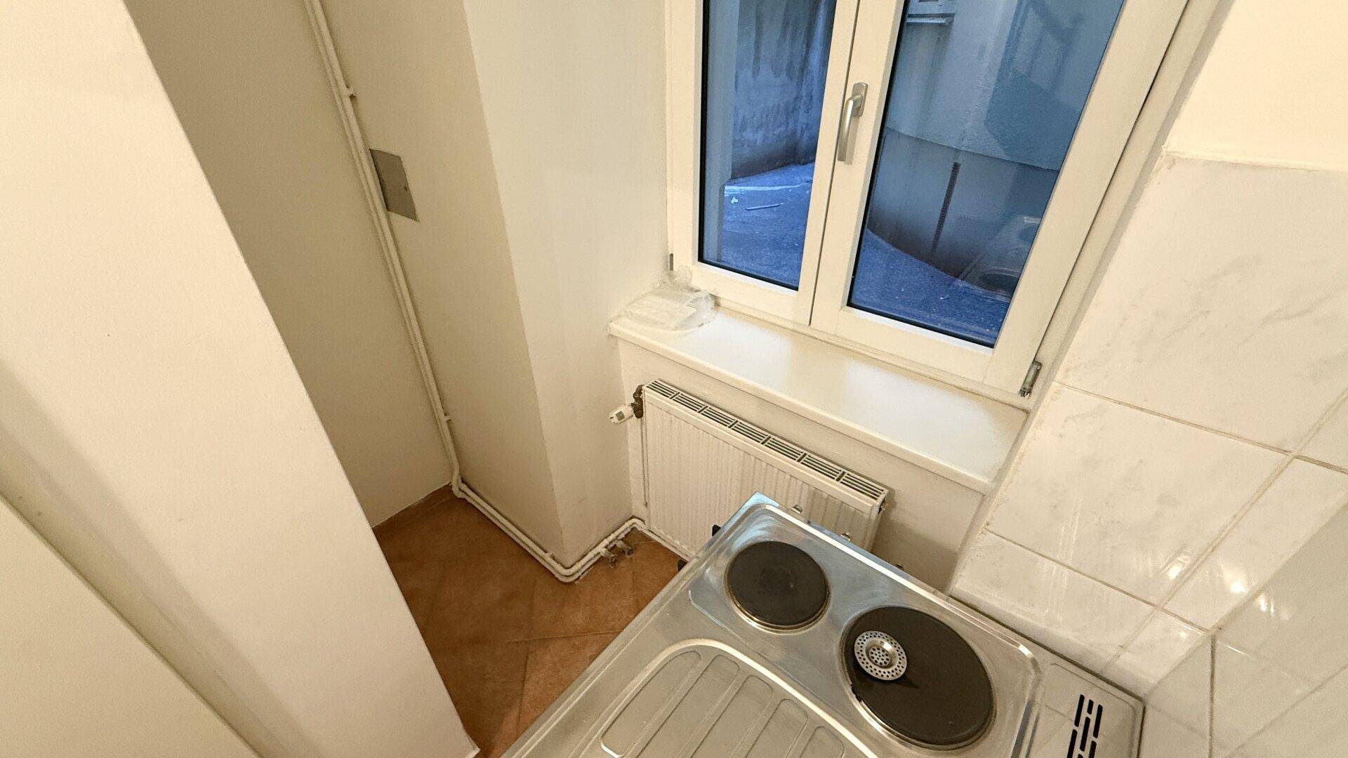 Kochnische mit Fenster, Spüle und Kochplatten, umgeben von hellen Fliesen und Wänden.