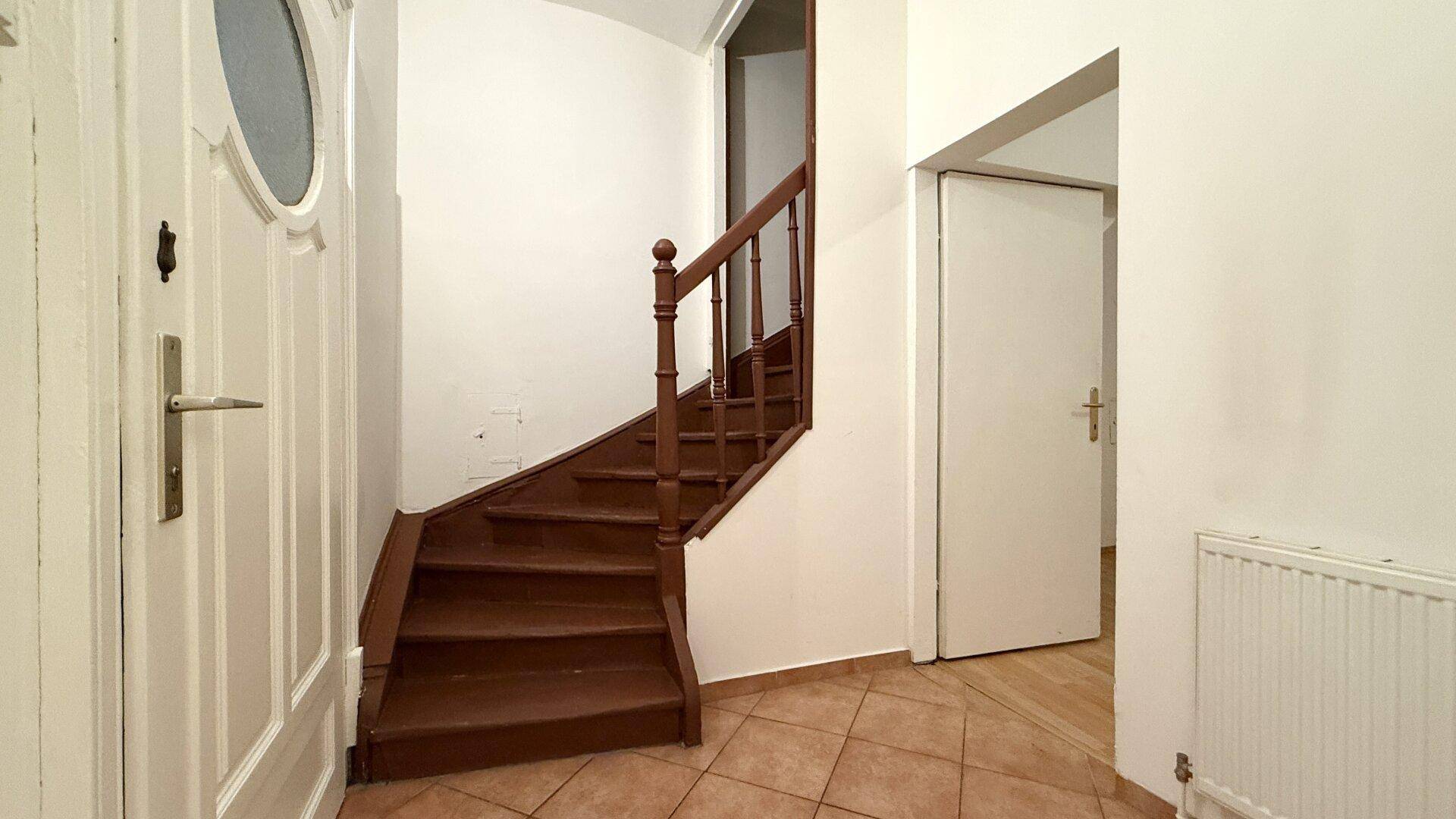 Eingangsbereich mit braunen Fliesen, einer Holztreppe und einer weißen Eingangstür.