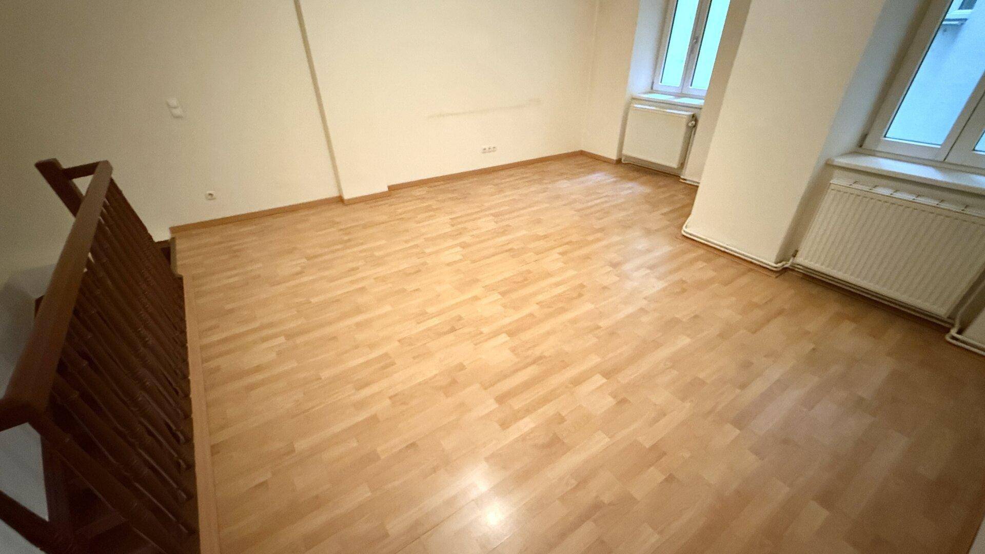 Heller Wohnraum mit Laminatboden und zwei Fenstern, die viel Tageslicht hereinlassen.
