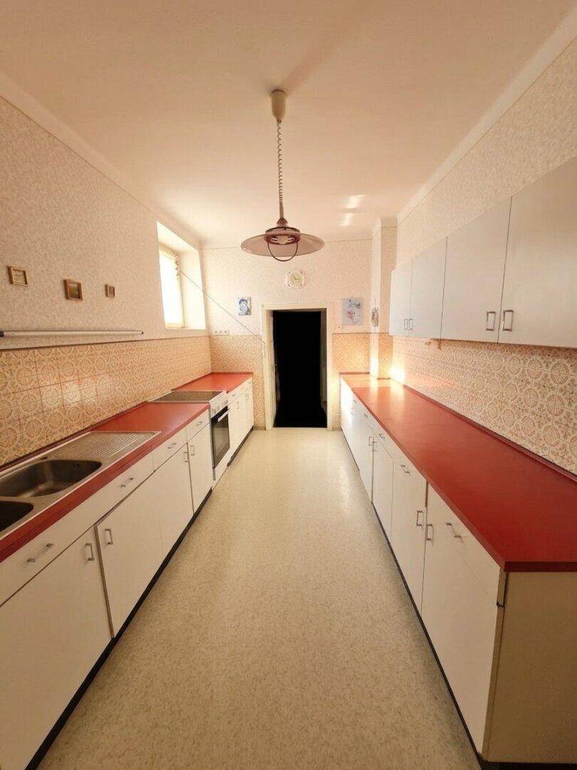 Schmale Küche mit weißen Schränken, roten Arbeitsplatten und Linoleumboden.