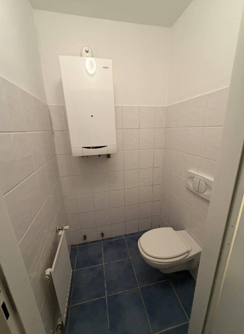 Separate Toilette mit weißen Wandfliesen, blauem Boden, wandhängendem WC und Warmwasserbereiter.