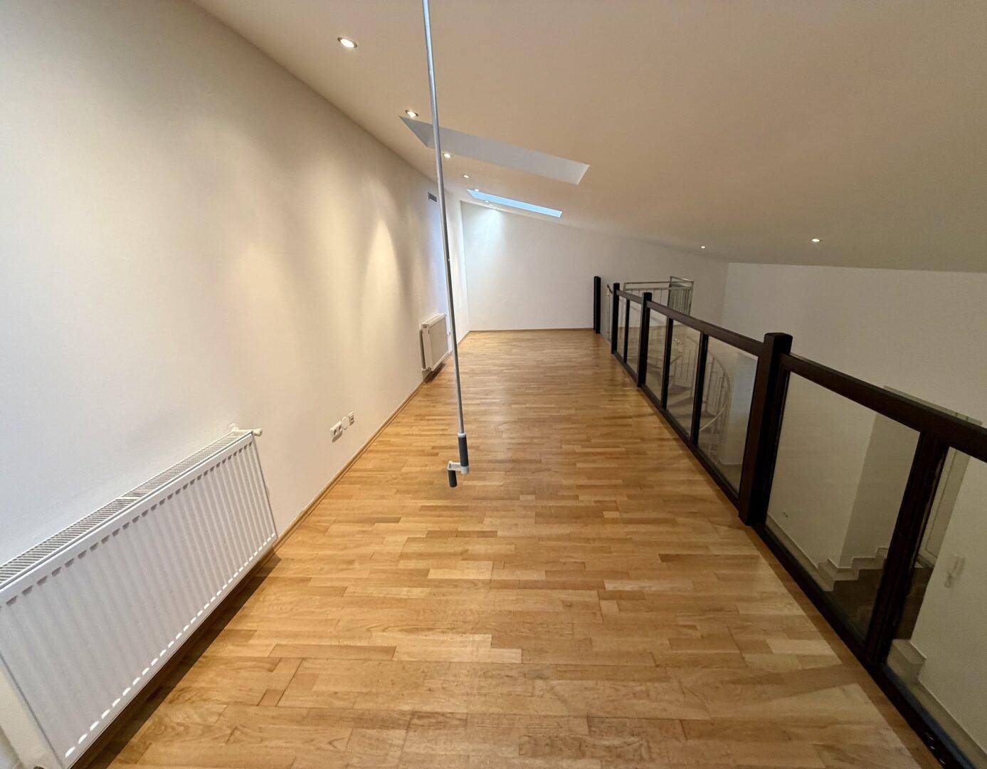 Offene Galerie mit hellem Holzfußboden, Glasgeländer und Dachfenstern, die viel Licht hereinlassen.