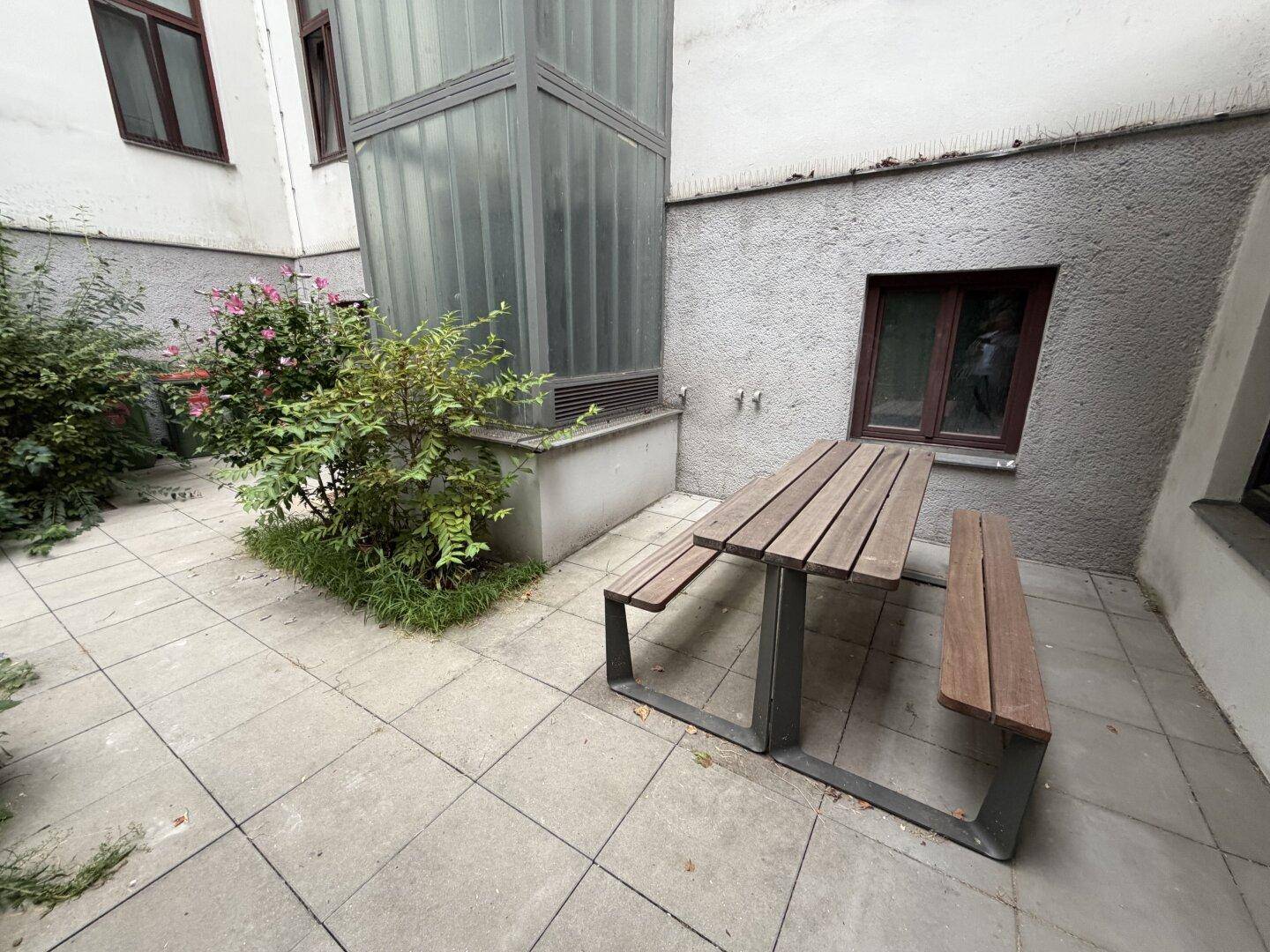 Gepflasterter Innenhof mit einem Holztisch, Bepflanzung und einem externen Aufzugsschacht.