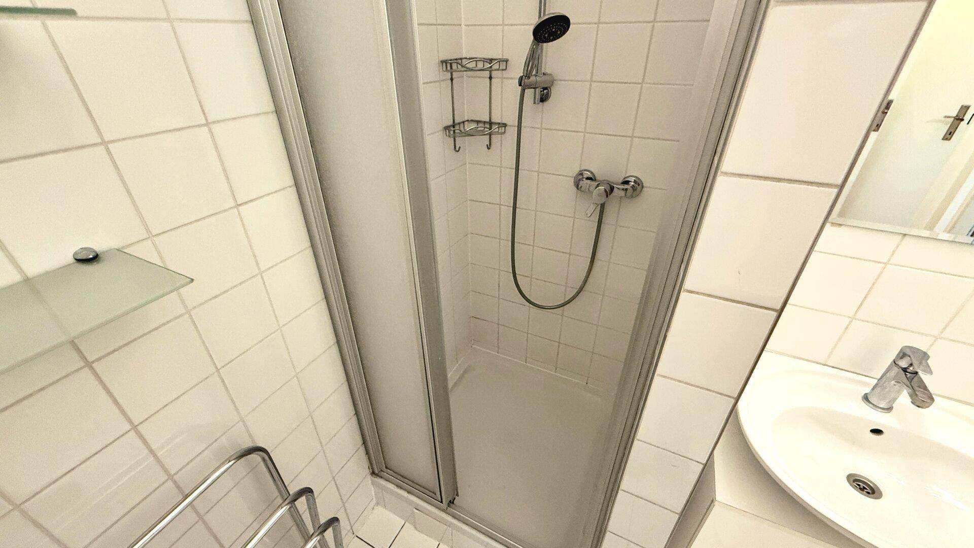 Duschkabine mit Glasabtrennung und Handbrause in einem weiß gefliesten Badezimmer.