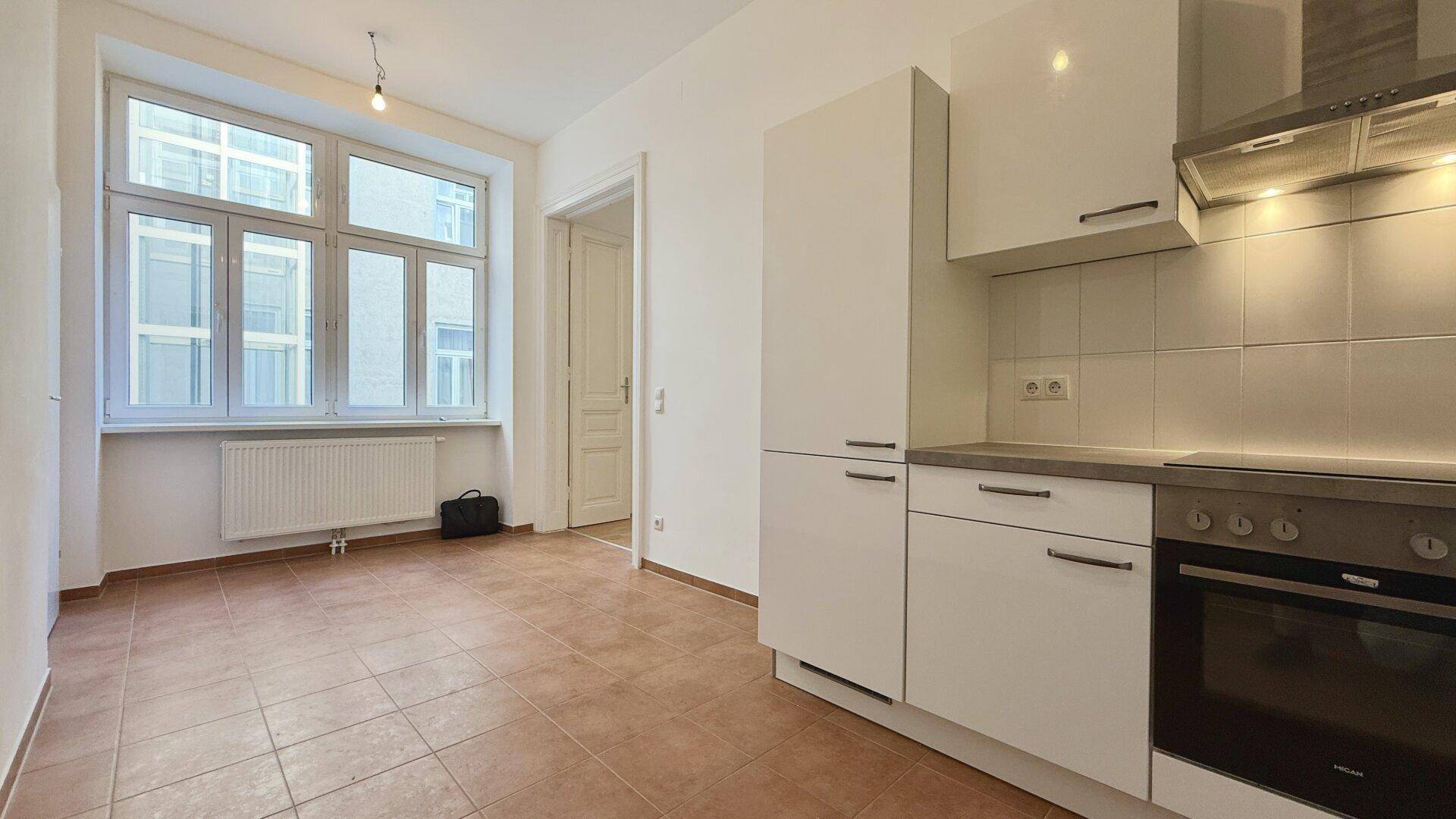 Helle Küche mit Terracotta-Fliesenboden und großem Fenster, angrenzend an einen weiteren Raum.