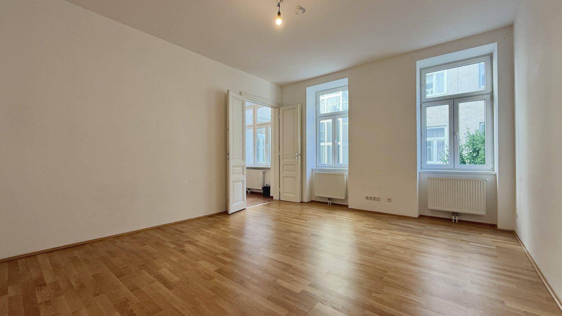 Lichtdurchflutetes Zimmer mit Parkettboden und hohen Decken, bietet Zugang zu einem weiteren Raum.