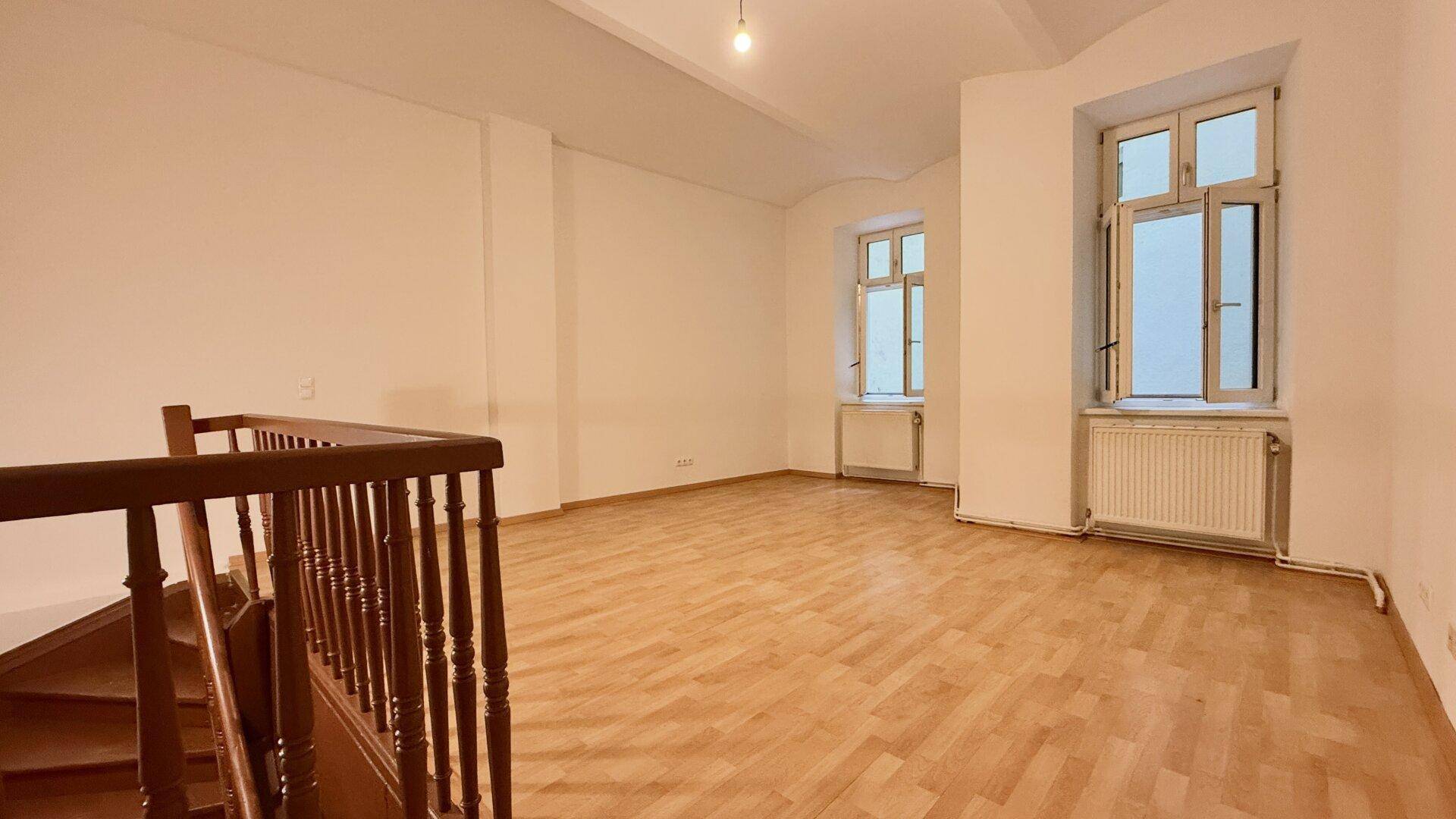 Geräumiger, leerer Raum mit Holzboden, hohen Fenstern und einem Treppengeländer.