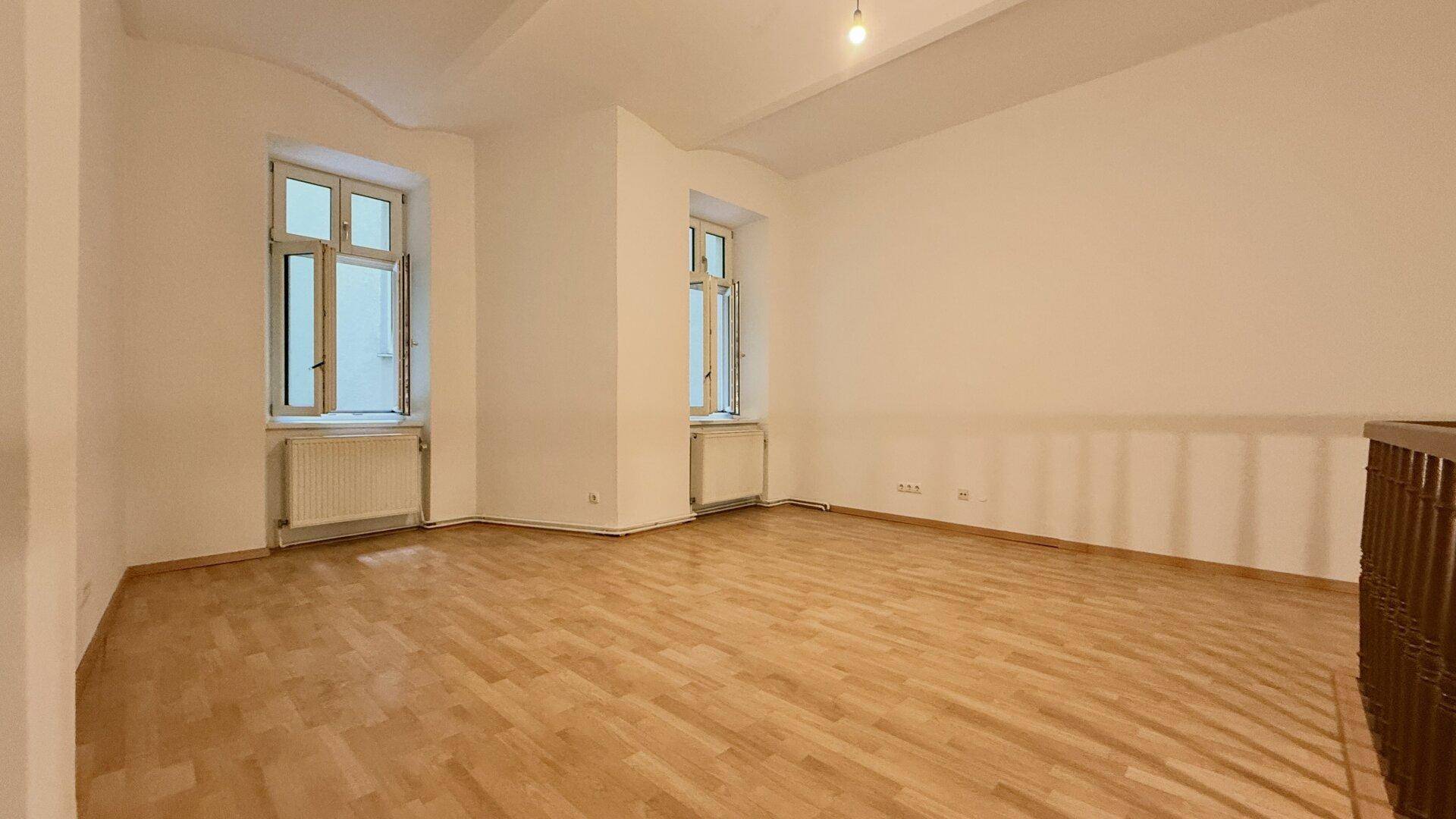 Großer, leerer Raum mit Holzboden und zwei hohen Fenstern, die viel Tageslicht hereinlassen.