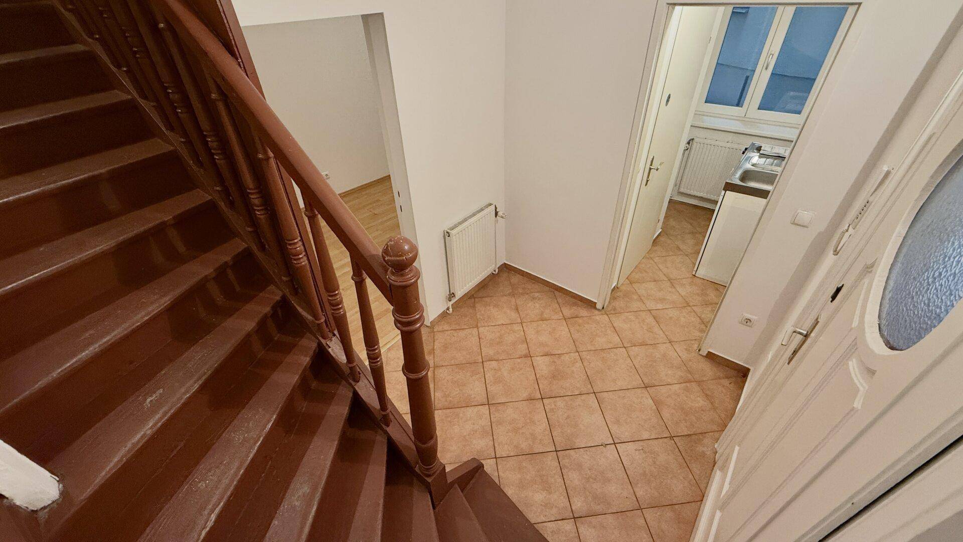 Blick von der Holztreppe auf den gefliesten Eingangsbereich mit Tür und kleiner Küchenzeile im Hintergrund.