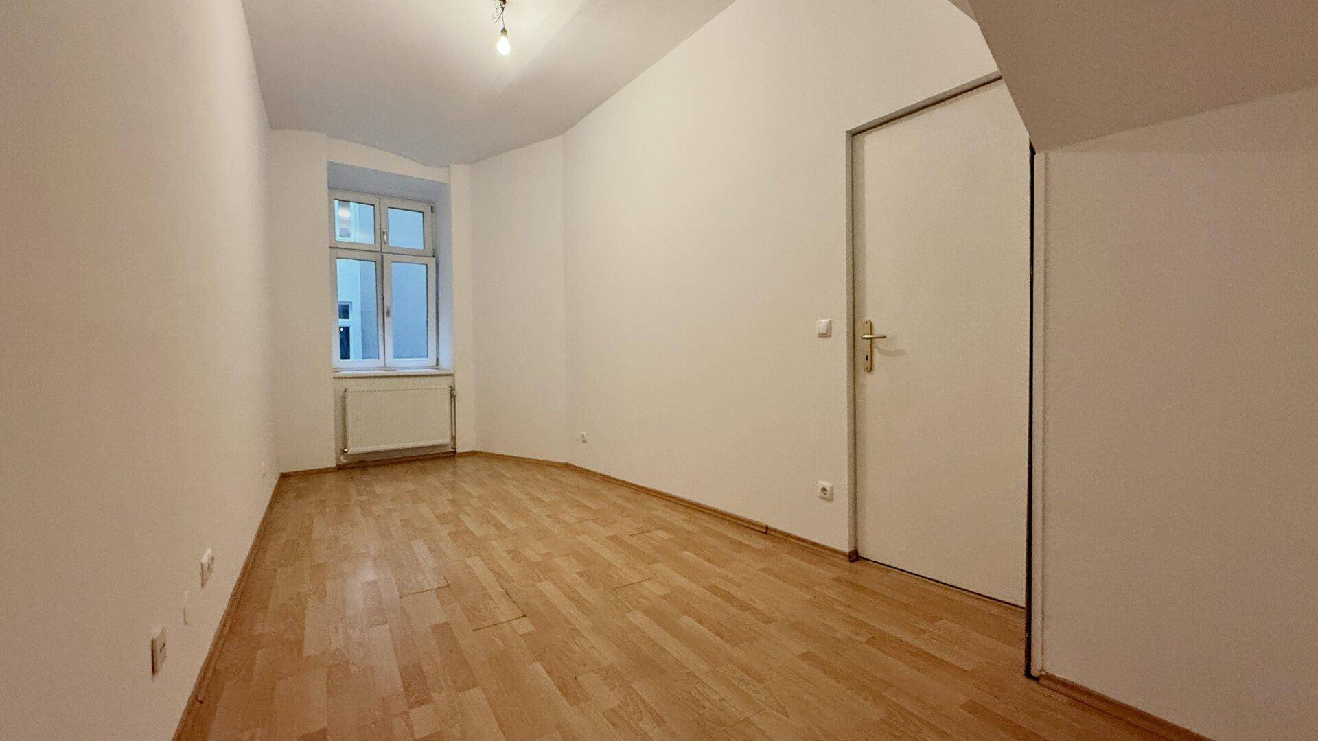 Leerer Raum mit Holzboden, weißen Wänden und einem Fenster, das Tageslicht hereinlässt.