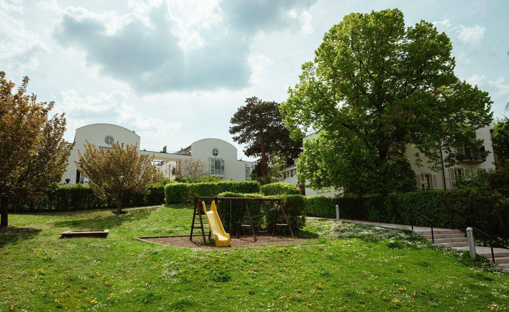 Grüne Gartenanlage mit Kinderspielplatz, umgeben von gepflegten Hecken und Bäumen.