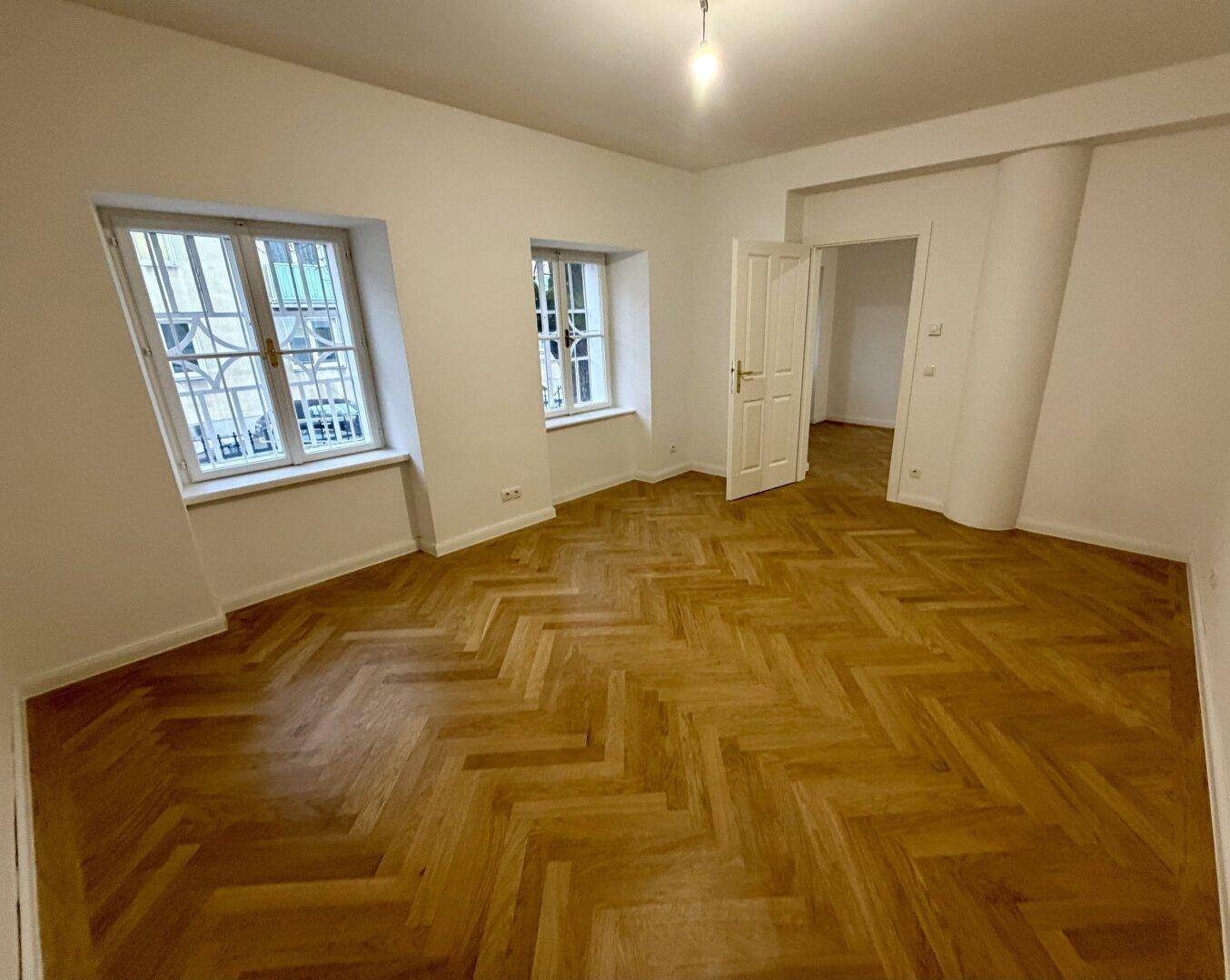 Geräumiges Wohnzimmer mit klassischem Fischgrätparkett und zwei Fenstern.