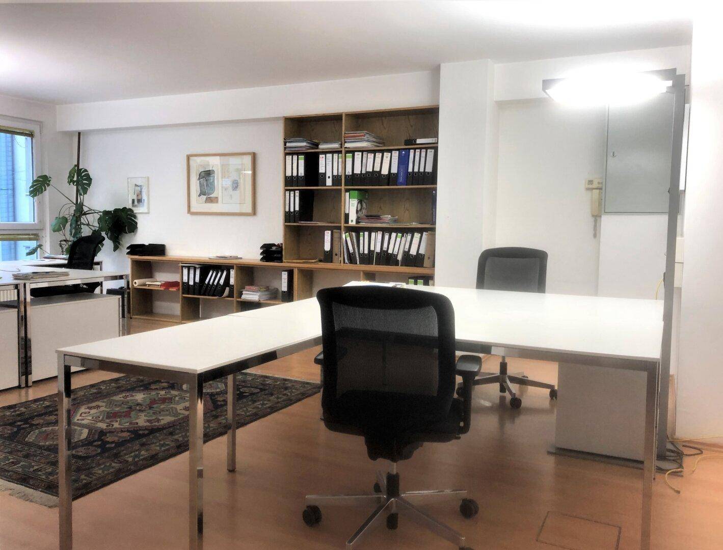 Büro mit mehreren Schreibtischen, Holzregalen und einem Teppich auf dem Holzfußboden, gut beleuchtet.