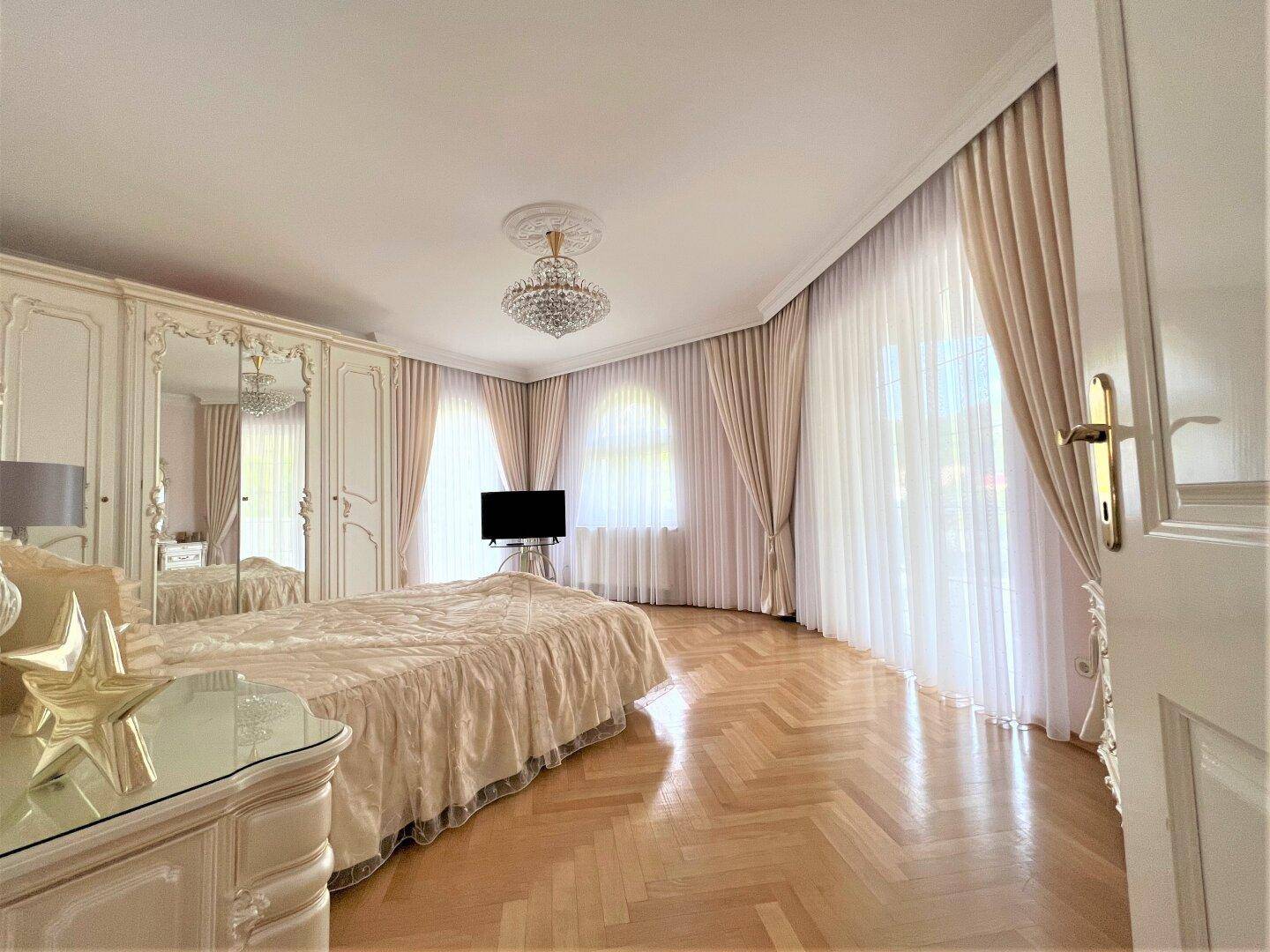 Helles Schlafzimmer mit elegantem Parkettboden, großen Fenstern und luxuriösem Kronleuchter.