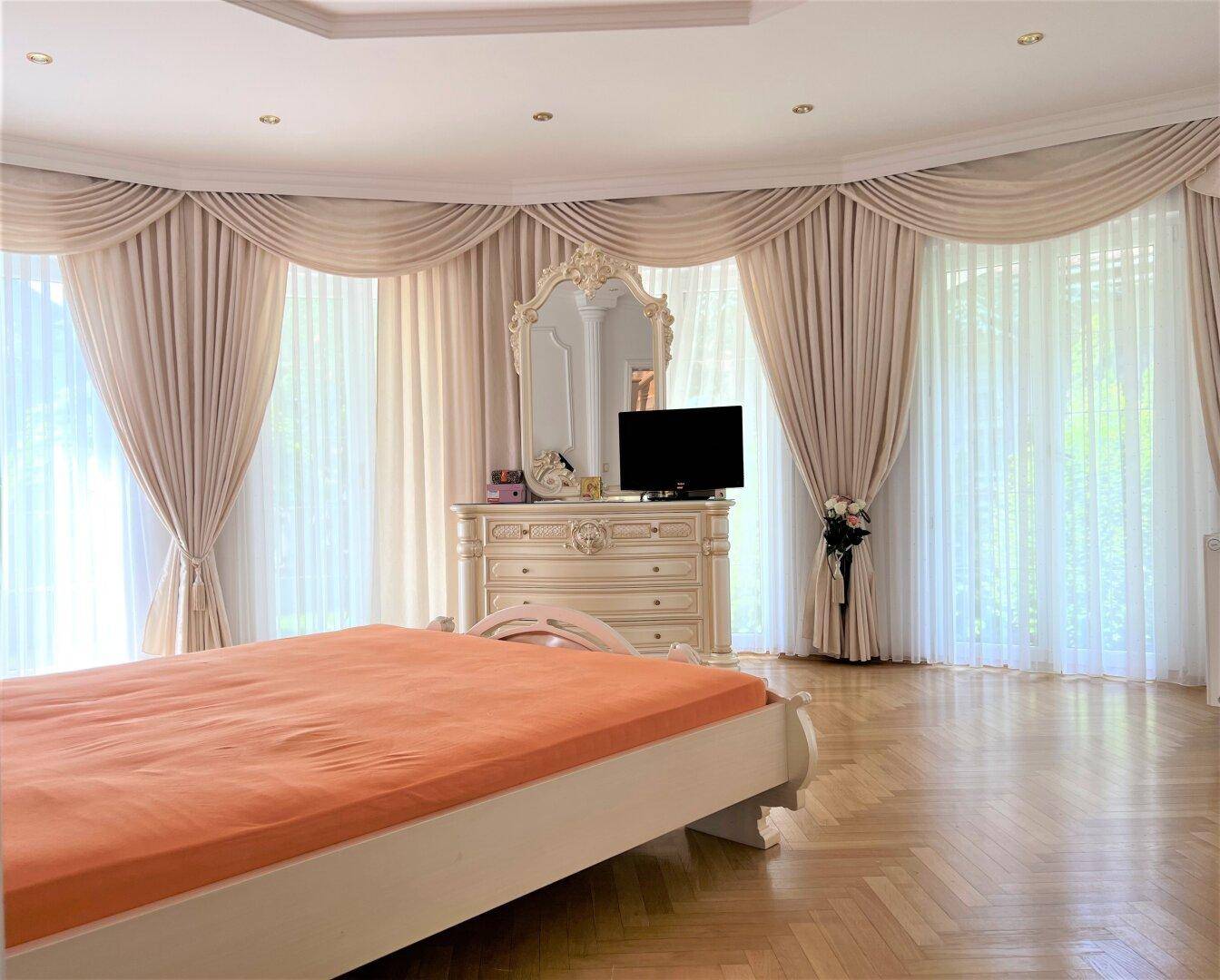 Helles Schlafzimmer mit großen Fenstern, Parkettboden und klassischer Einrichtung.