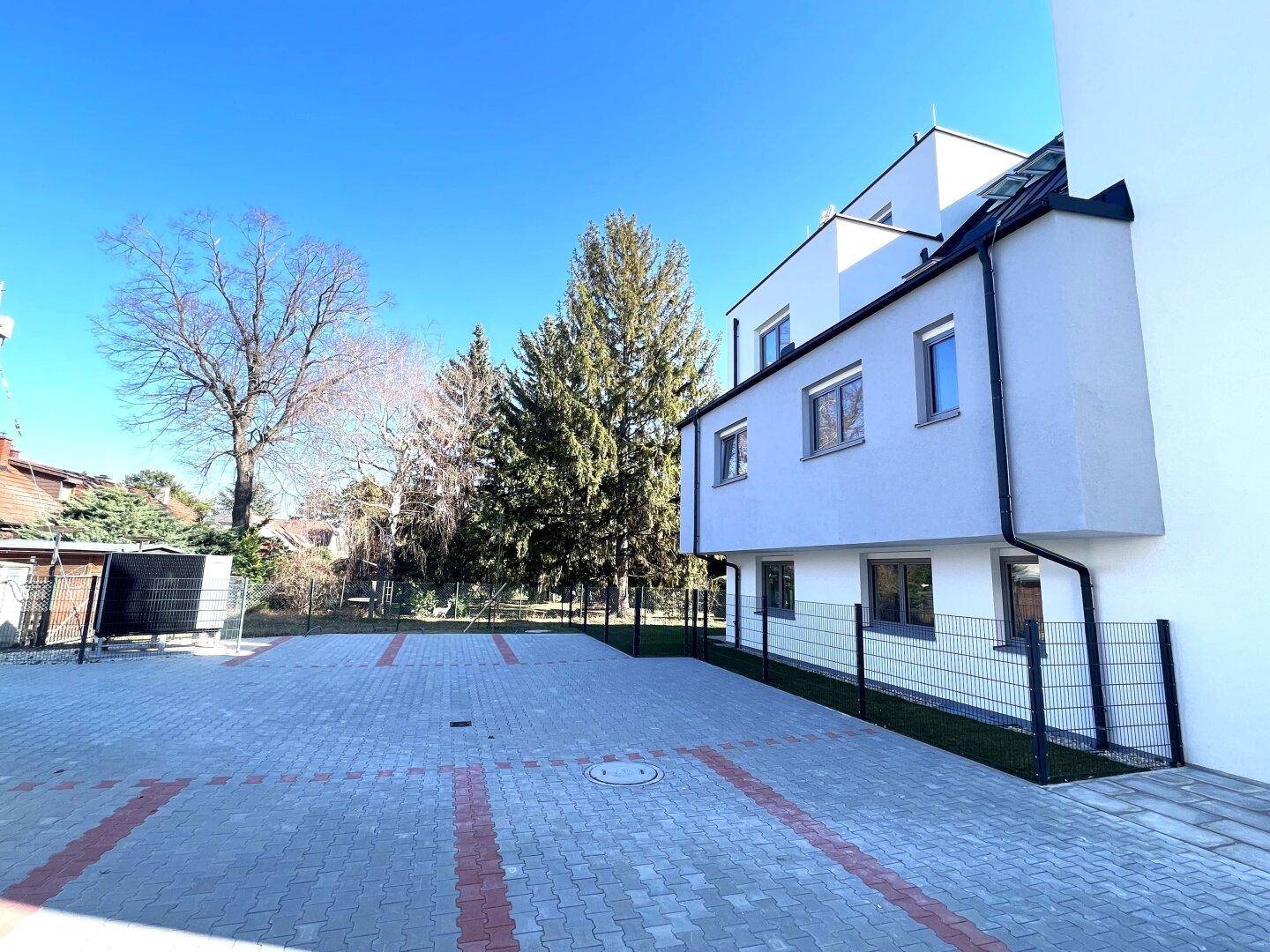 Außenansicht des Neubaus mit großem gepflasterten Parkplatz und angrenzendem Grünbereich.