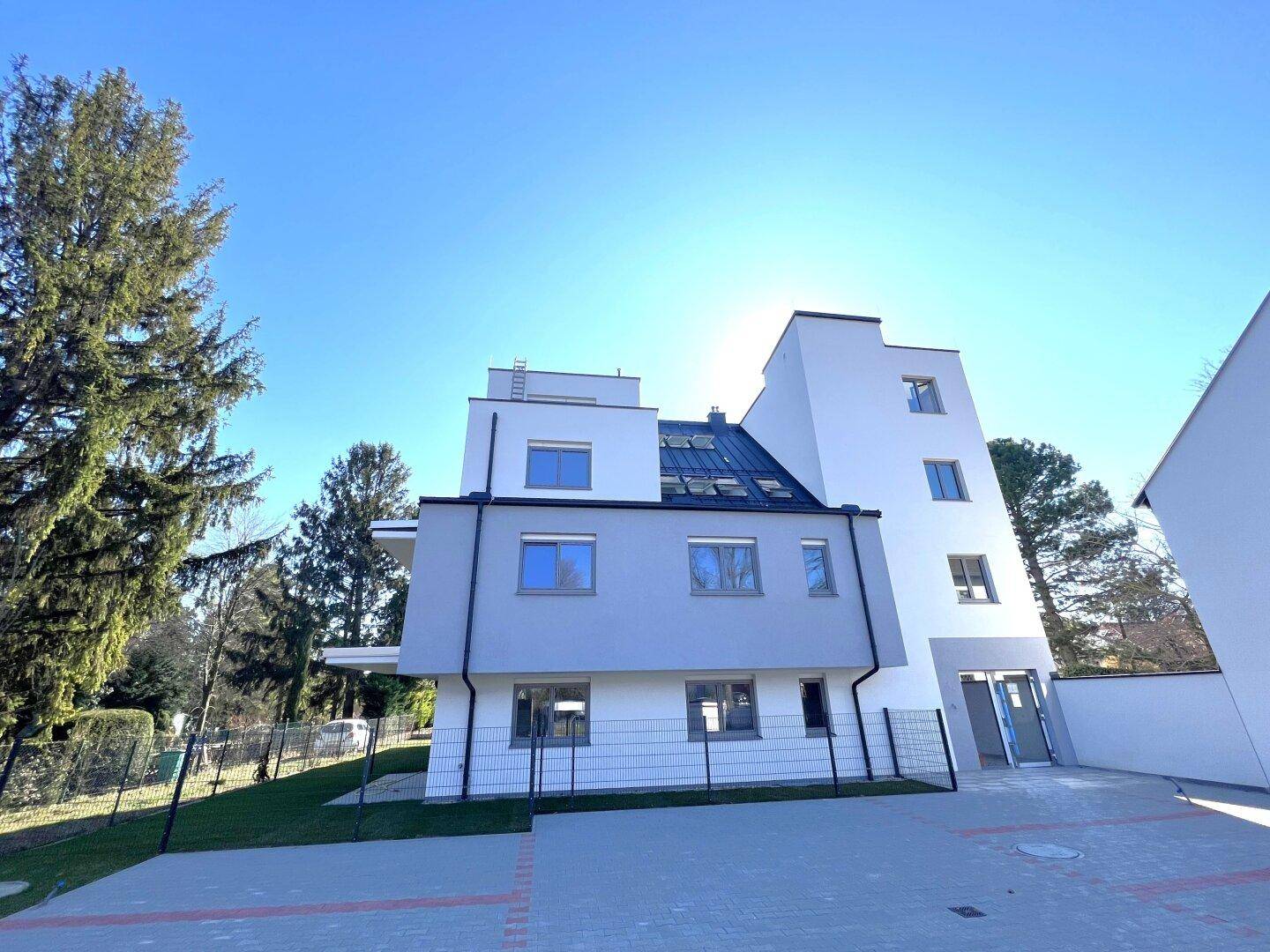 Neubaugebäude mit moderner Architektur, großem Parkplatz und grüner Umgebung bei Sonnenschein.