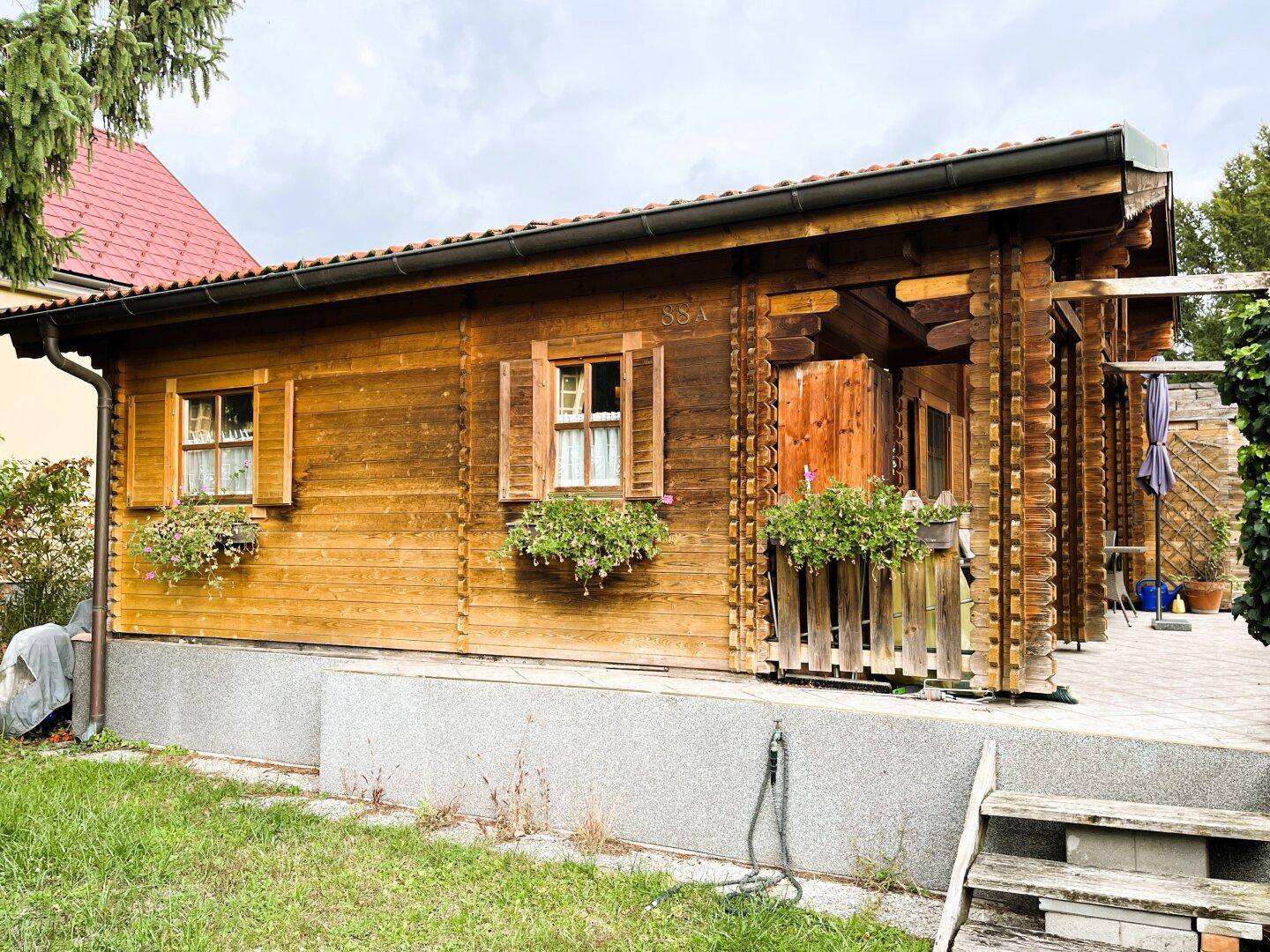 Seitenansicht des Holzhauses mit traditionellen Fensterläden und einem kleinen Gartenbereich.