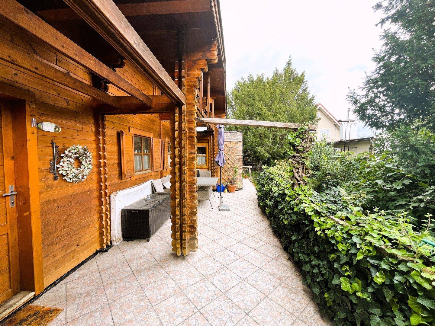 Lange, geflieste Terrasse entlang des Holzhauses mit Sitzgelegenheiten und üppiger Bepflanzung.