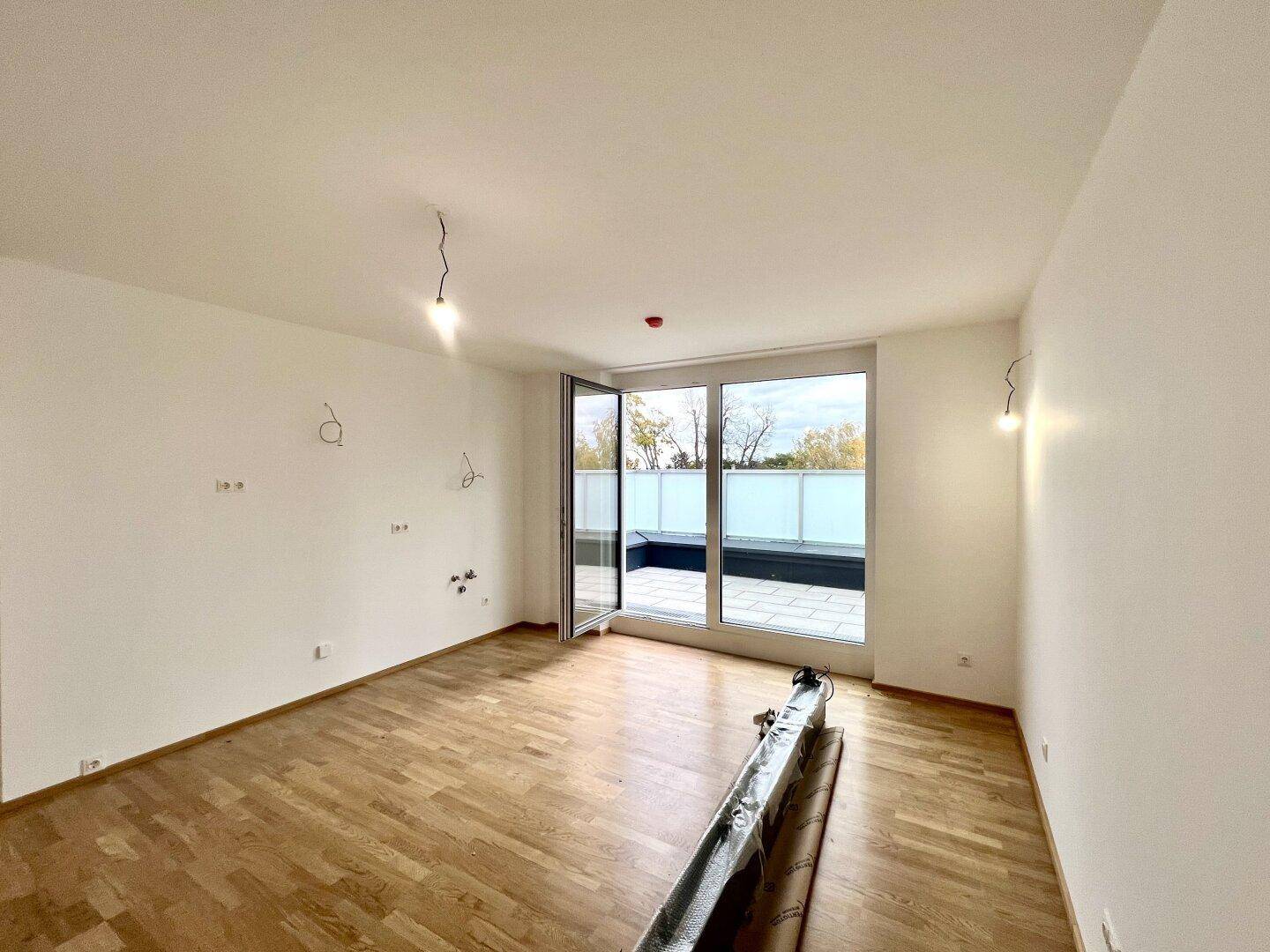Heller, unmöblierter Raum mit Holzboden und großen Fenstern, die auf eine Terrasse führen.