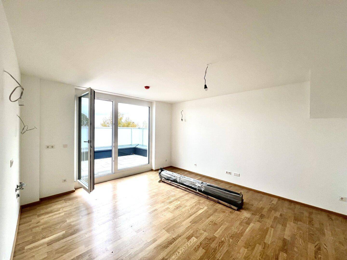 Unmöblierter Raum mit Holzboden und großen Fenstern, die den Zugang zu einer Terrasse ermöglichen.