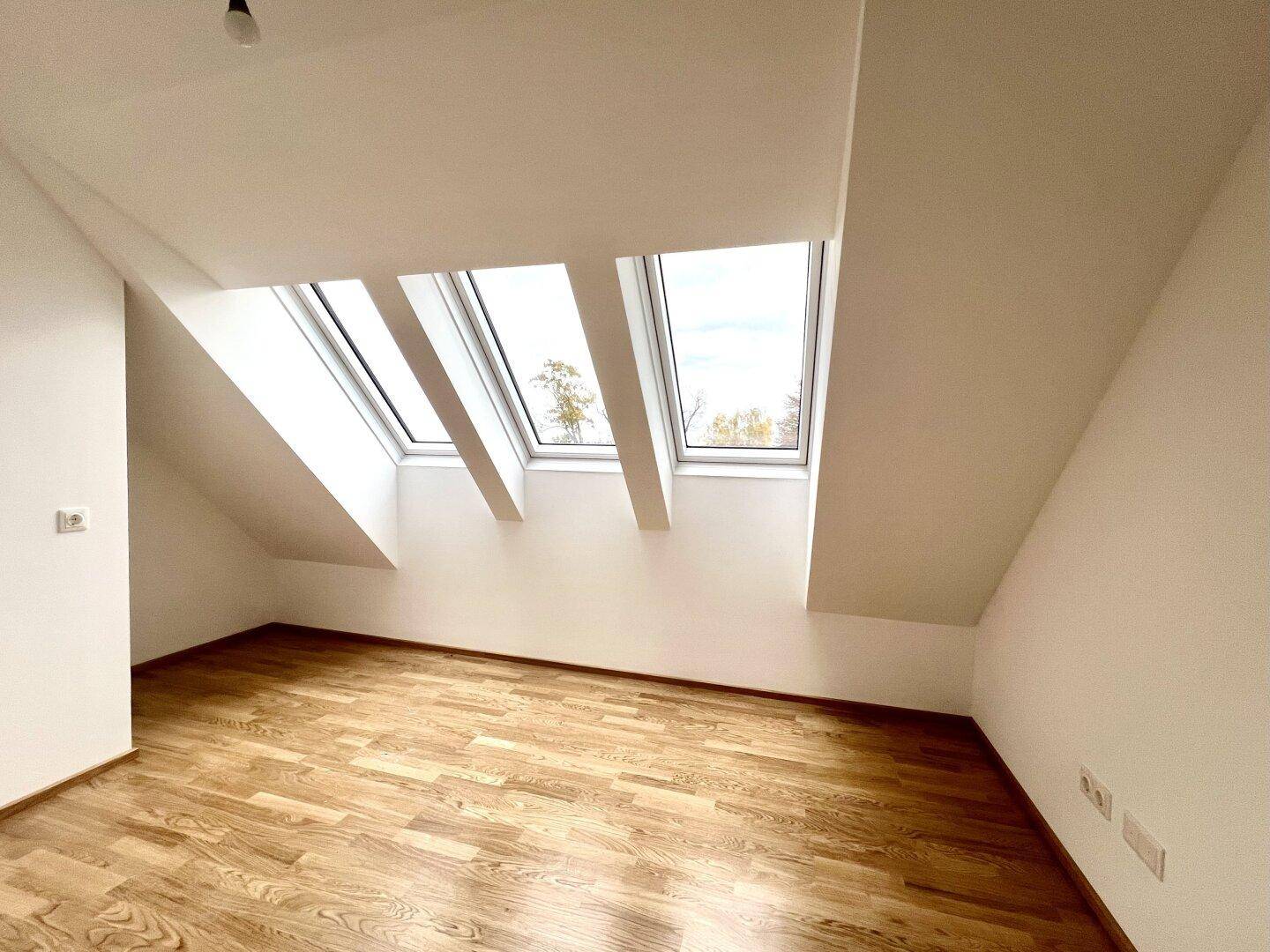 Helles Dachzimmer mit Holzboden und mehreren Dachfenstern, die viel Tageslicht hereinlassen.