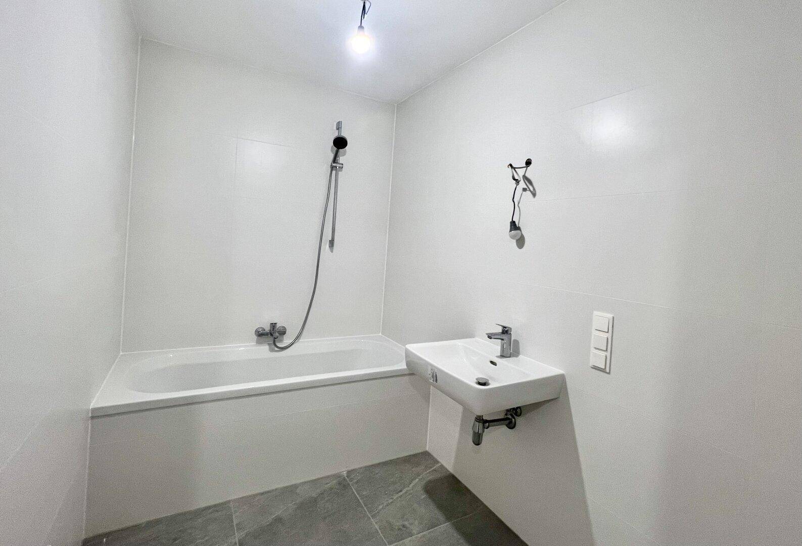 Modernes Badezimmer mit Badewanne, Waschbecken und grauen Fliesen, in neuwertigem Zustand.