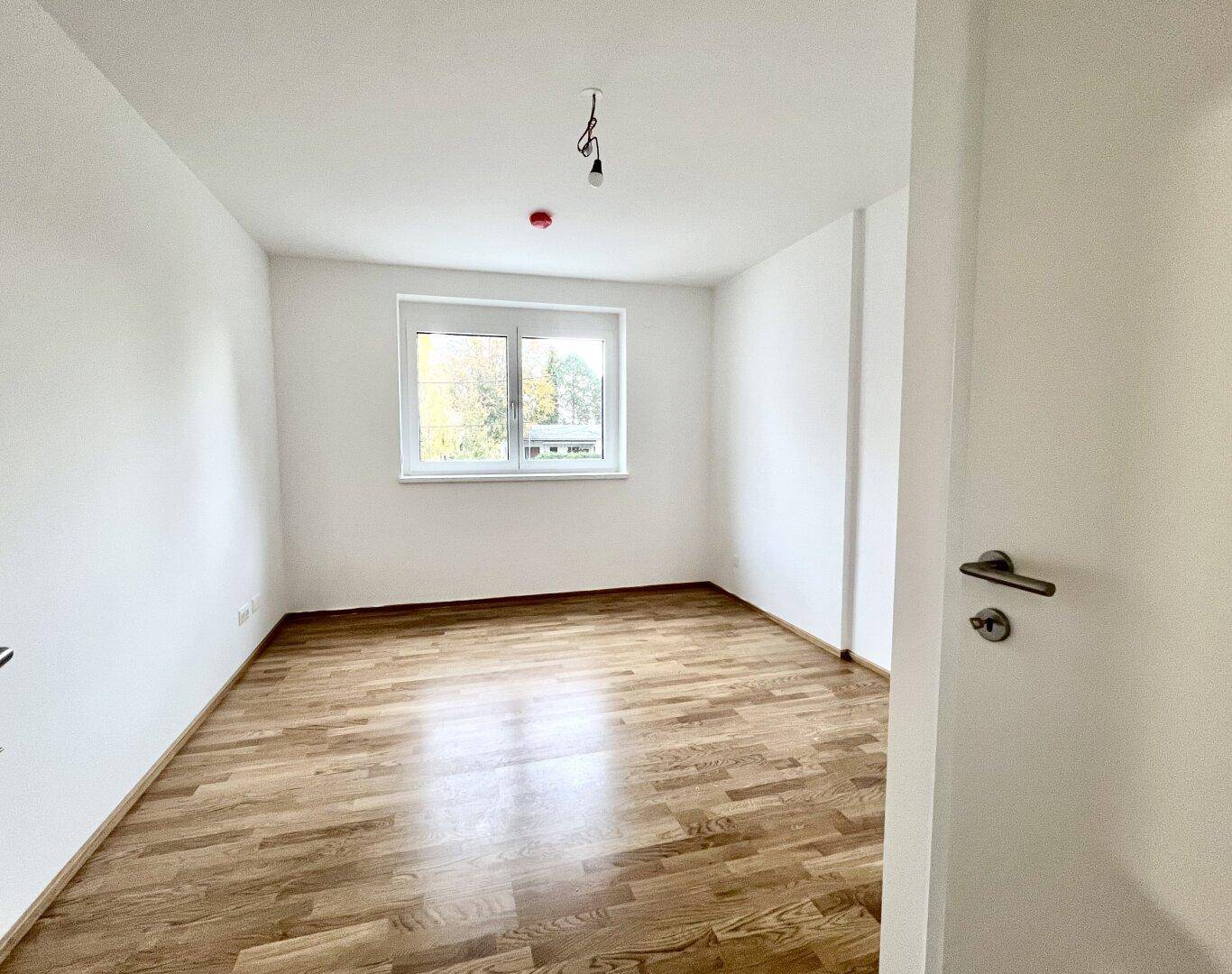 Helles Schlafzimmer mit Parkettboden und einem Fenster, das den Blick ins Grüne freigibt.