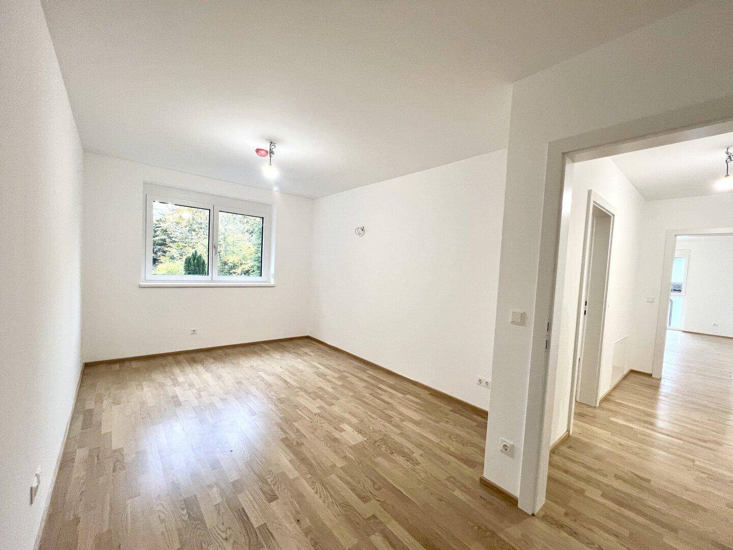 Geräumiger, heller Raum mit Holzboden, weißen Wänden und Blick ins Grüne durch ein großes Fenster.