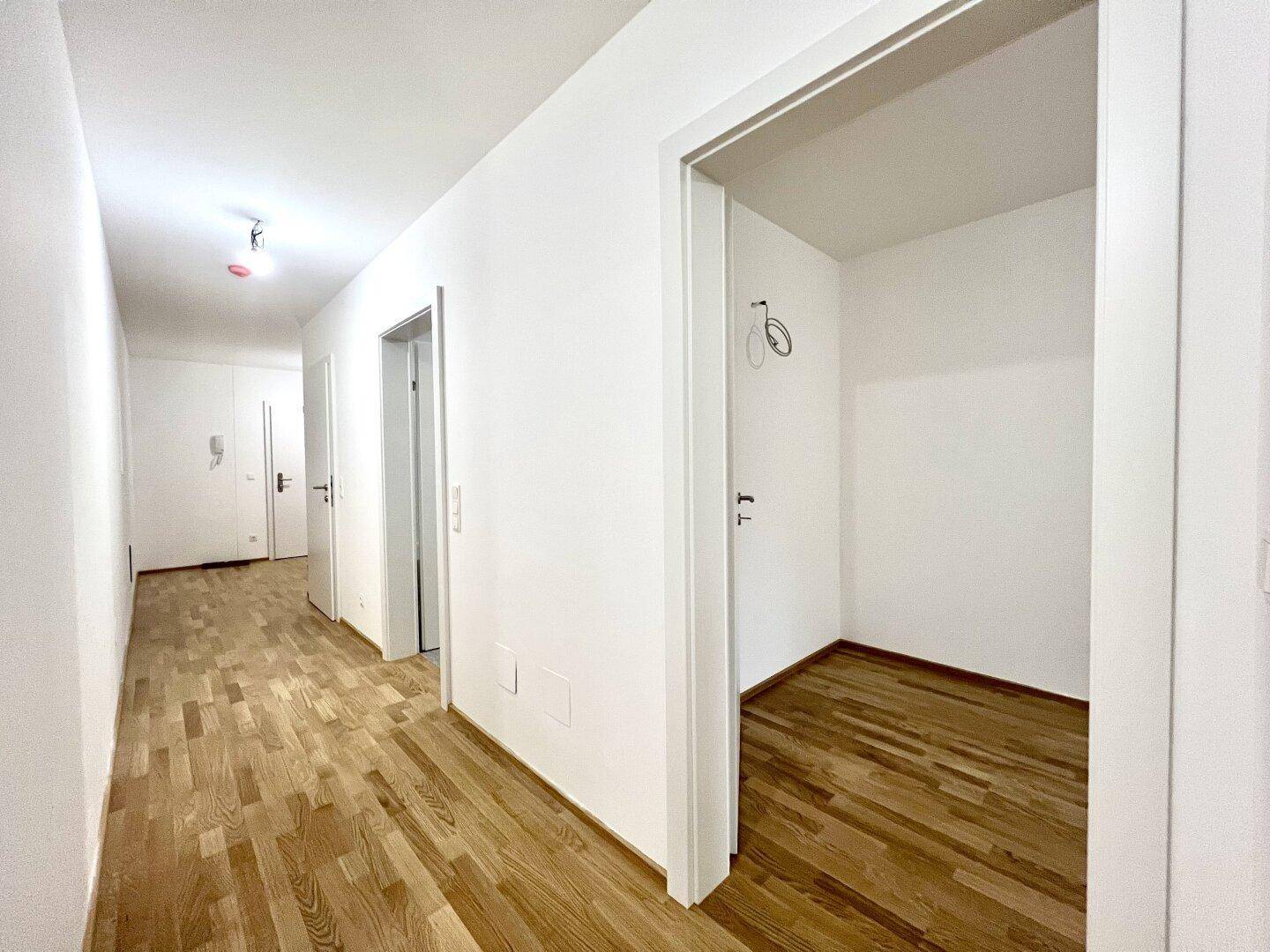 Langer Flur mit Holzboden und weißen Wänden, der zu verschiedenen Räumen und einem Abstellbereich führt.